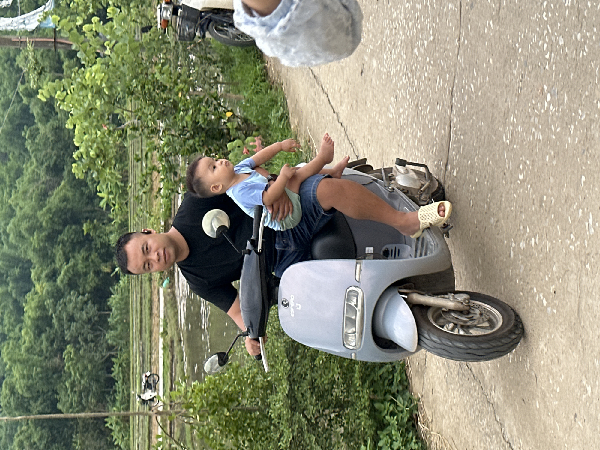 In Chiềng Châu, Vietnam, a man is riding a light blue electric scooter on a paved road. He is wearing a black t-shirt and denim shorts, and a young child, dressed in a light blue t-shirt and green shorts, is seated on his lap. The child is facing to the left, and the man's left arm is around the child. The scooter has a prominent front headlight and is partially obscured by the rider and passenger. In the background, there are lush green trees, a body of water with reflections, and terraced fields that are characteristic of the region. Another scooter is visible further down the road to the left. The overall scene depicts a common mode of transportation and family life in a rural Vietnamese setting.