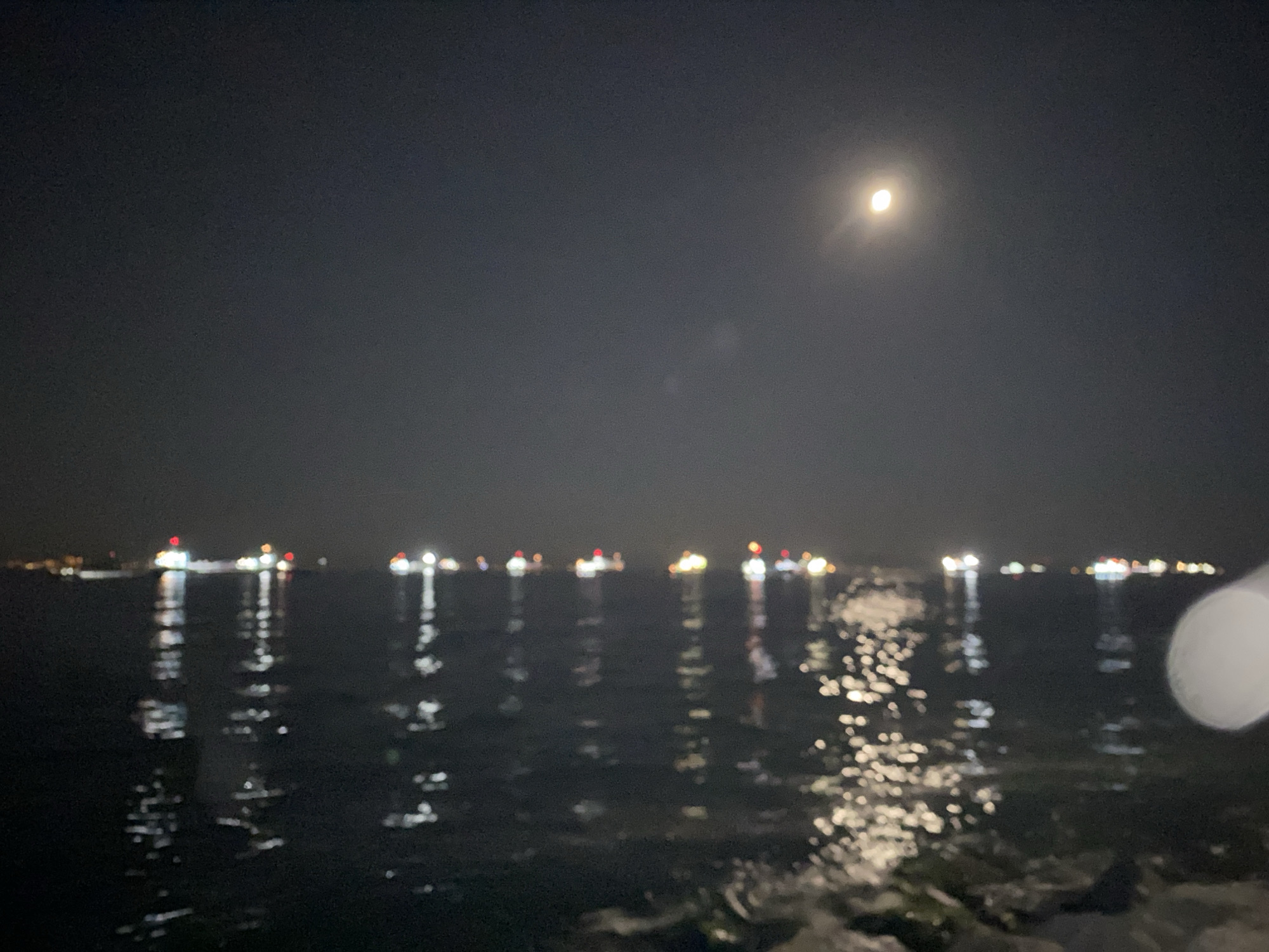 This is a nighttime, out-of-focus photograph of the Istanbul skyline at sea. In the distance, a line of ships with their lights on are visible, their reflections shimmering on the water. The moon is visible in the upper right quadrant of the sky. The foreground shows the dark, choppy water with rocks along the edge of what appears to be a pier or shoreline. The overall impression is one of a serene, slightly mysterious night scene.