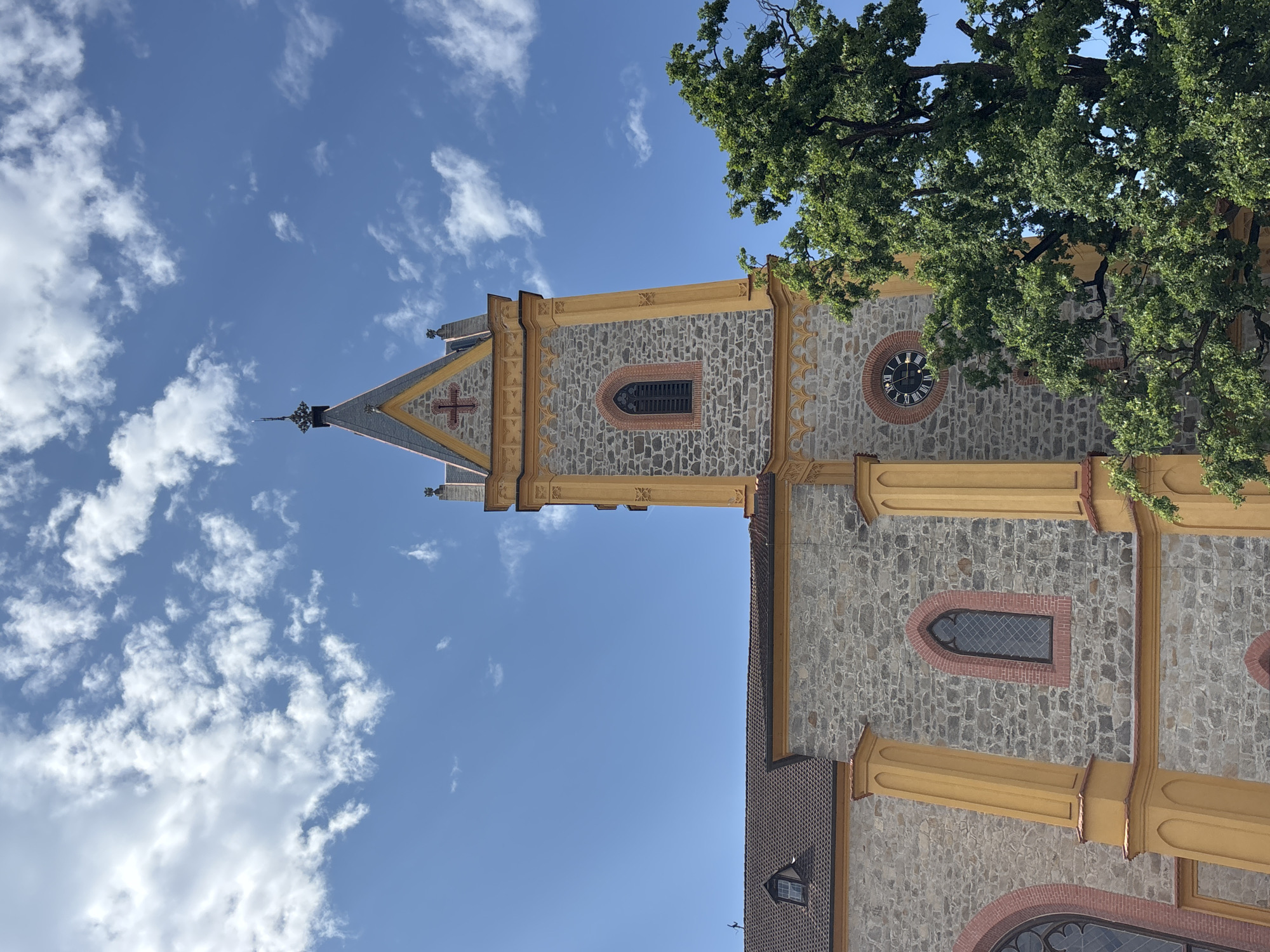 The image shows the tower and a section of the facade of the Hluboká nad Vltavou church, located in Hluboká nad Vltavou, Czechia. The architecture is characterized by stone walls with yellow decorative elements and arched windows. The church tower is tall and slender, topped with a pointed roof and a cross. A clock face is visible on the tower. Lush green trees are in the foreground, partially obscuring the church building. The sky is blue with scattered white clouds. There are no people visible in the image.