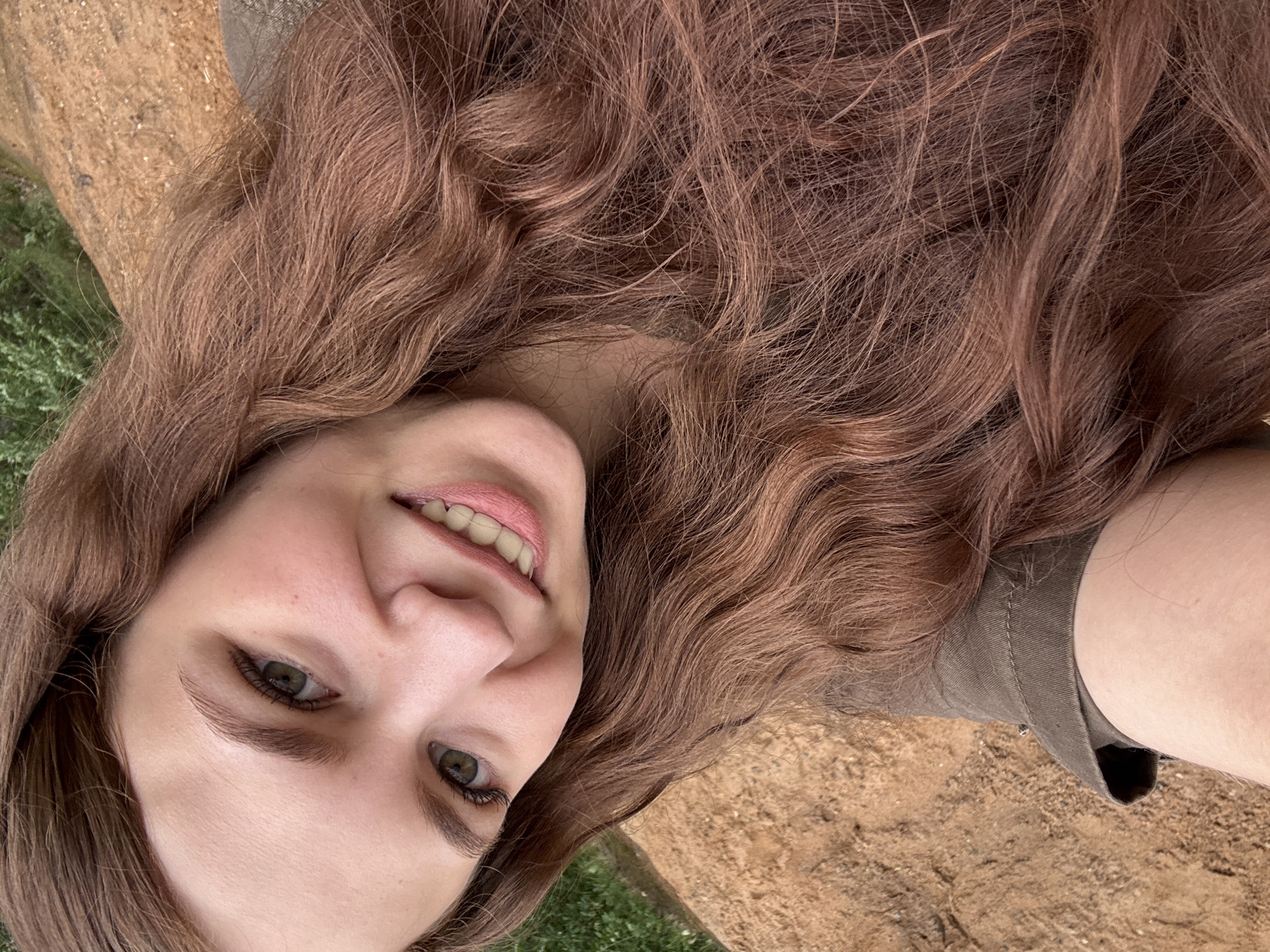 A young woman with long, wavy, reddish-brown hair is captured in a close-up, low-angle selfie. She is outdoors, with a blurred background of green foliage and sandy, earthy terrain. Her face is turned towards the camera, and she has a bright, open-mouthed smile, revealing straight, white teeth. Her eyes are a striking green, and her eyebrows are well-defined. She appears to be wearing a khaki-colored jacket. The lighting suggests it is daytime. There is no information in the image that would suggest a location within the city of Chaikovsky, Russia.