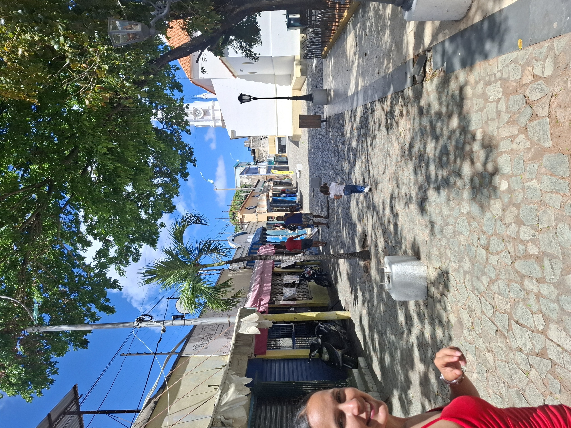 This image depicts a sunny street scene in Juan Griego, Venezuela, captured from a low angle, partially as a selfie. In the bottom right foreground, the smiling face and red clothing of the person taking the photograph are visible.
The street, paved with irregular stone tiles, stretches away from the viewer, dappled with sharp shadows cast by trees and buildings, indicating strong daylight. Several people populate the scene: one individual in a white shirt and blue jeans bends over in the mid-ground, while two others stand further back near shops—one in a blue top and shorts, another in a red top. More figures are discernible in the distance.
Along the right side of the street stands a prominent white building with a red tiled roof and a clock tower, suggesting a church or significant public building. A classic black street lamp is positioned nearby. On the left, a row of single-story shops features colorful awnings in pink, yellow, and blue, with motorcycles parked in front of some. Utility poles and a network of wires are visible overhead on the left.
Lush green foliage from a large tree occupies the upper left foreground, with a decorative lantern hanging from its branches. A slender palm tree stands further down the street on the left. The sky is a brilliant, clear blue with a few fluffy white clouds, underscoring the bright, sunny weather. A grey concrete block or planter is situated on the right side of the paved area.
Visible text on a shop sign includes "PLAZA *DEL*" and "Tienda" (Spanish for "Shop"). The scene conveys a relaxed, everyday atmosphere in a vibrant town, likely during mid-morning or early afternoon due to the intense sunlight and distinct shadows.