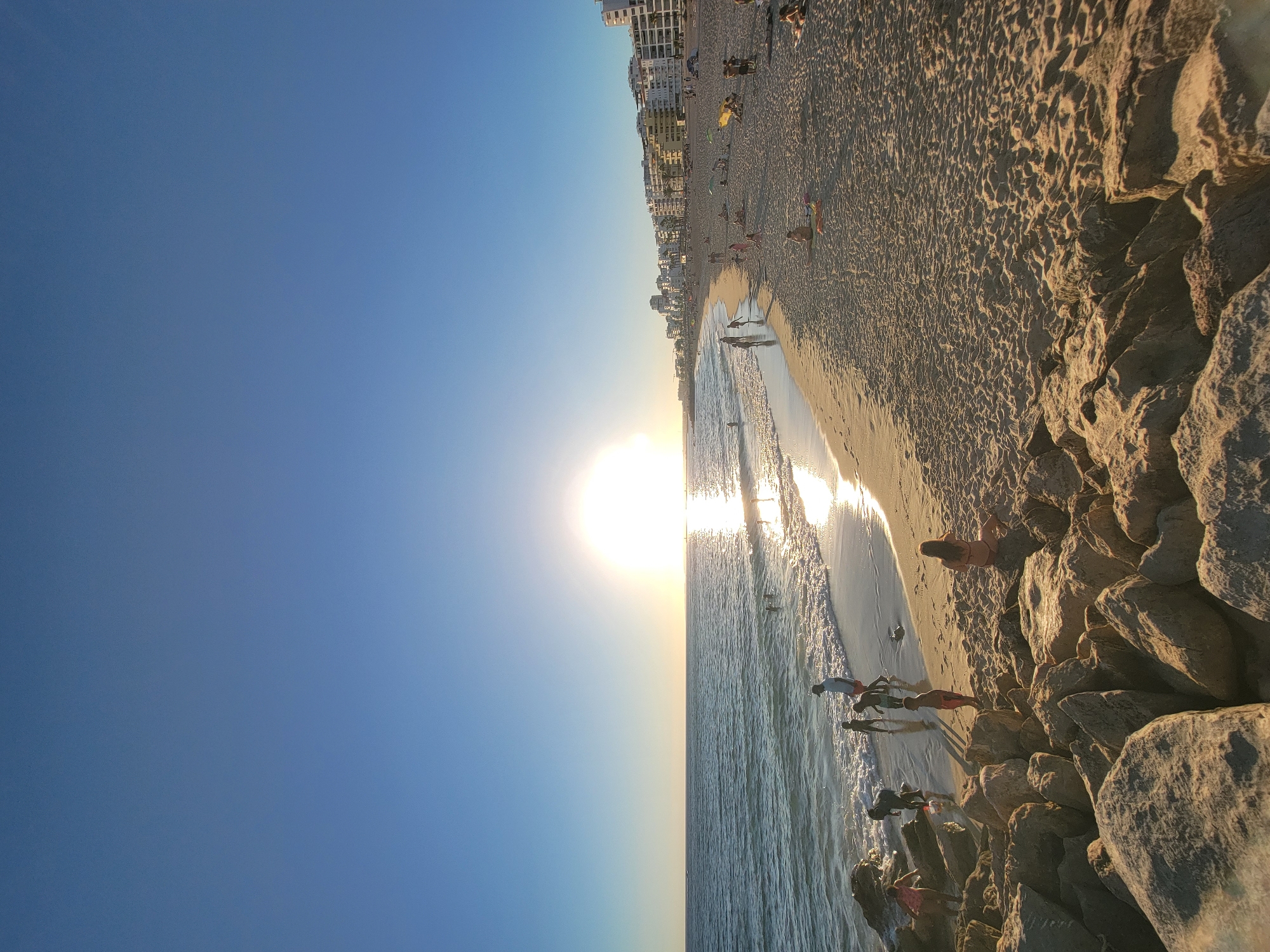 This image captures a serene beach scene in Quarteira, Portugal, during what appears to be a late afternoon or early evening, given the low angle of the sun. The sun is a prominent feature, positioned over the ocean, casting a bright, shimmering reflection on the water's surface and bathing the beach in a warm, golden light.
The foreground is dominated by a rocky shoreline, with large, weathered stones creating a natural barrier. Beyond the rocks, the sandy beach stretches out, dotted with numerous people engaged in various activities. Some individuals are wading in the gentle waves lapping at the shore, while others are scattered across the sand, relaxing, playing, or strolling. Children are visible interacting in the shallow water and on the beach.
In the background, a strip of buildings, characteristic of a coastal resort town, lines the edge of the beach. These structures are a mix of white and cream-colored, with balconies and windows facing the sea. The sky is a clear, vibrant blue, transitioning to a lighter hue near the horizon where it meets the ocean. The overall impression is one of a peaceful and idyllic seaside setting, with the natural beauty of the coast complemented by the presence of human activity.