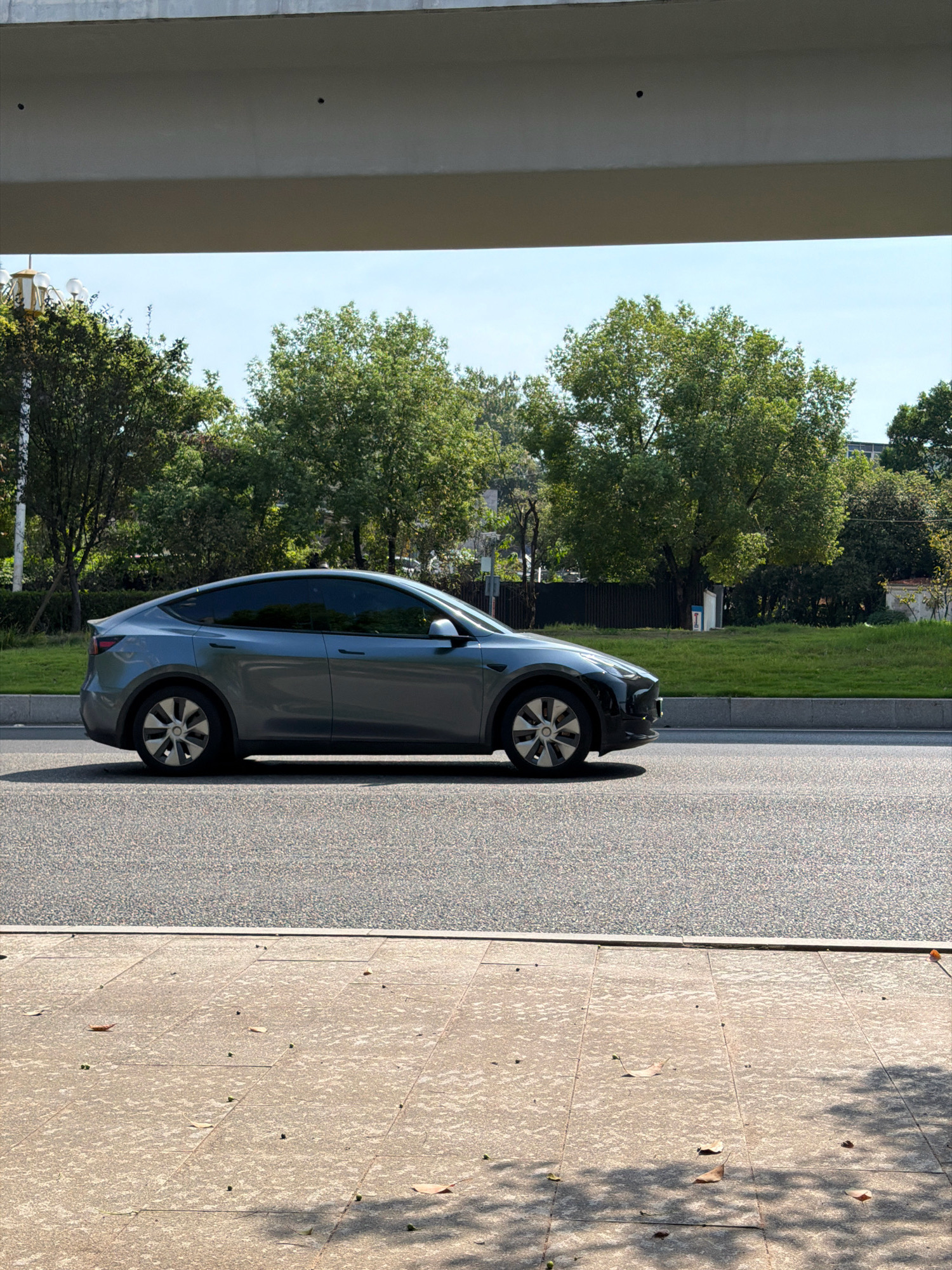 A grey Tesla Model Y is parked on the side of a road in Jiangjin, China, under an overpass. The car is positioned parallel to the road and facing right. The road is paved with asphalt. To the left of the road is a sidewalk with scattered fallen leaves. Beyond the road, there is a strip of green grass and then a line of trees. The sky is clear and blue. The overpass casts a shadow over the scene. The image is taken from a slightly elevated perspective, looking down at the car.