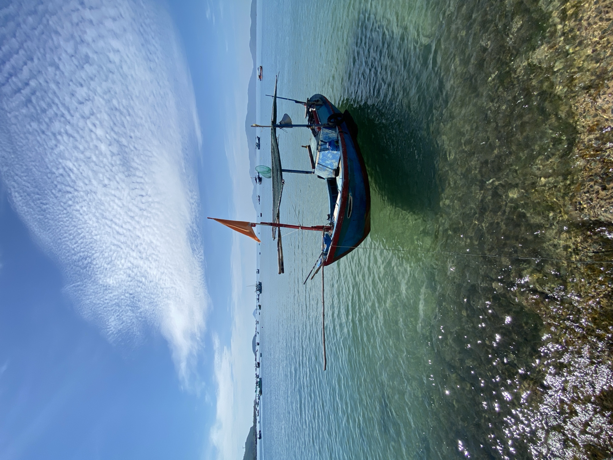 An anchored, blue and red fishing boat with an orange flag and a tarped canopy floats in the clear, shallow water of Nha Trang Bay, Vietnam, on a sunny day. The boat is positioned in the foreground, slightly right of center, with its bow facing left. The seabed is visible beneath the boat, with rocks and shadows casting into the water. In the background, a calm sea stretches to the horizon, dotted with numerous other fishing boats of varying sizes. Distant, hazy mountains form the backdrop. The sky above is a vibrant blue, with scattered, wispy white clouds. The image captures a serene, coastal scene, likely in the early morning or late afternoon given the gentle sunlight reflecting on the water.