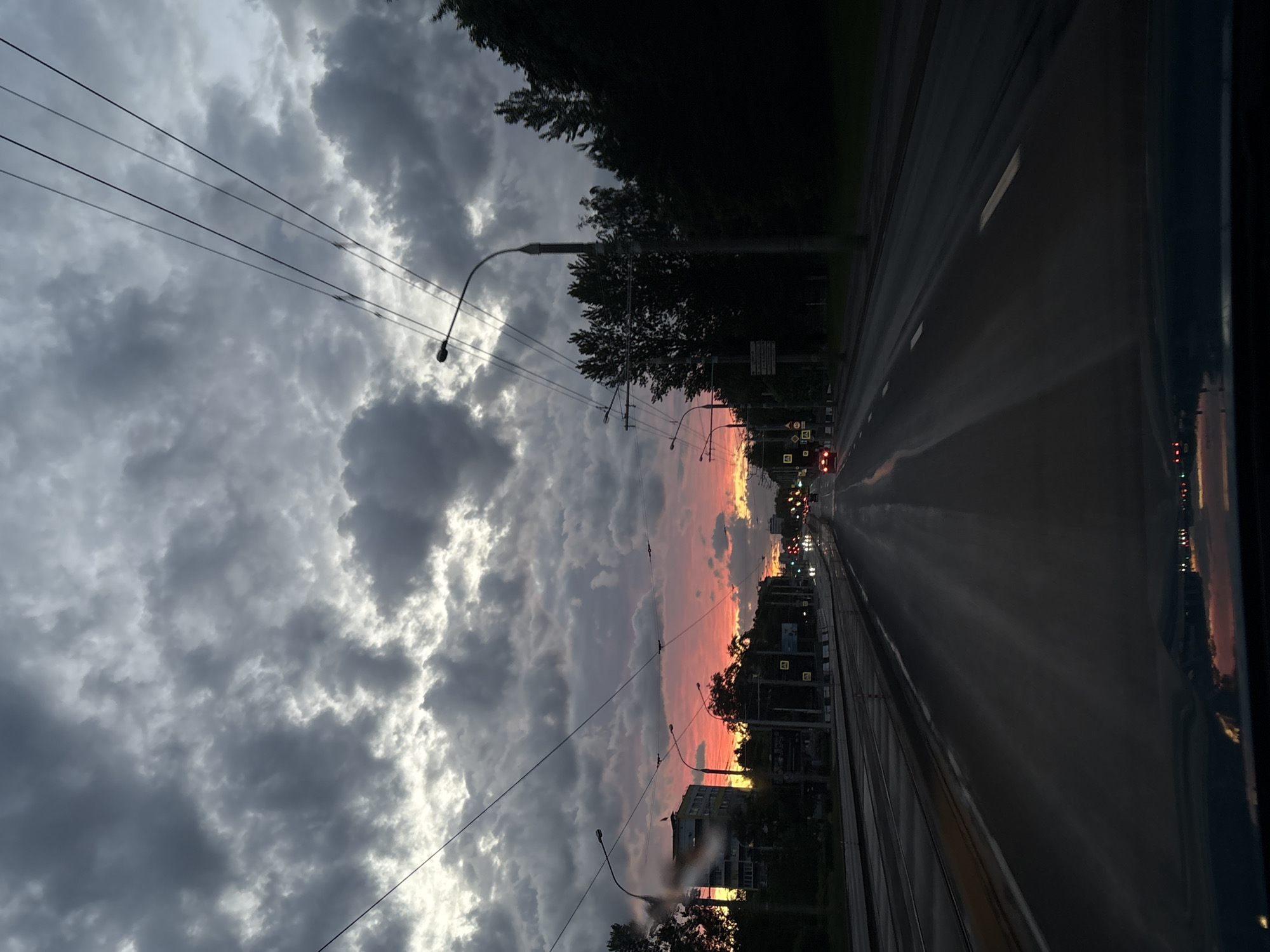 The image shows a view from a vehicle on a street in Minsk, Belarus, at sunset. The street is a multi-lane road with tram tracks running down the center. Streetlights line the sides of the road. Buildings are visible in the background, silhouetted against the sunset. The sky is filled with dramatic clouds, with the sun setting in the distance, casting a warm, orange glow. Overhead power lines are visible. Traffic lights are red in the distance. The street appears wet, reflecting the lights of the surrounding environment. The interior of the vehicle is visible on the right side of the image.