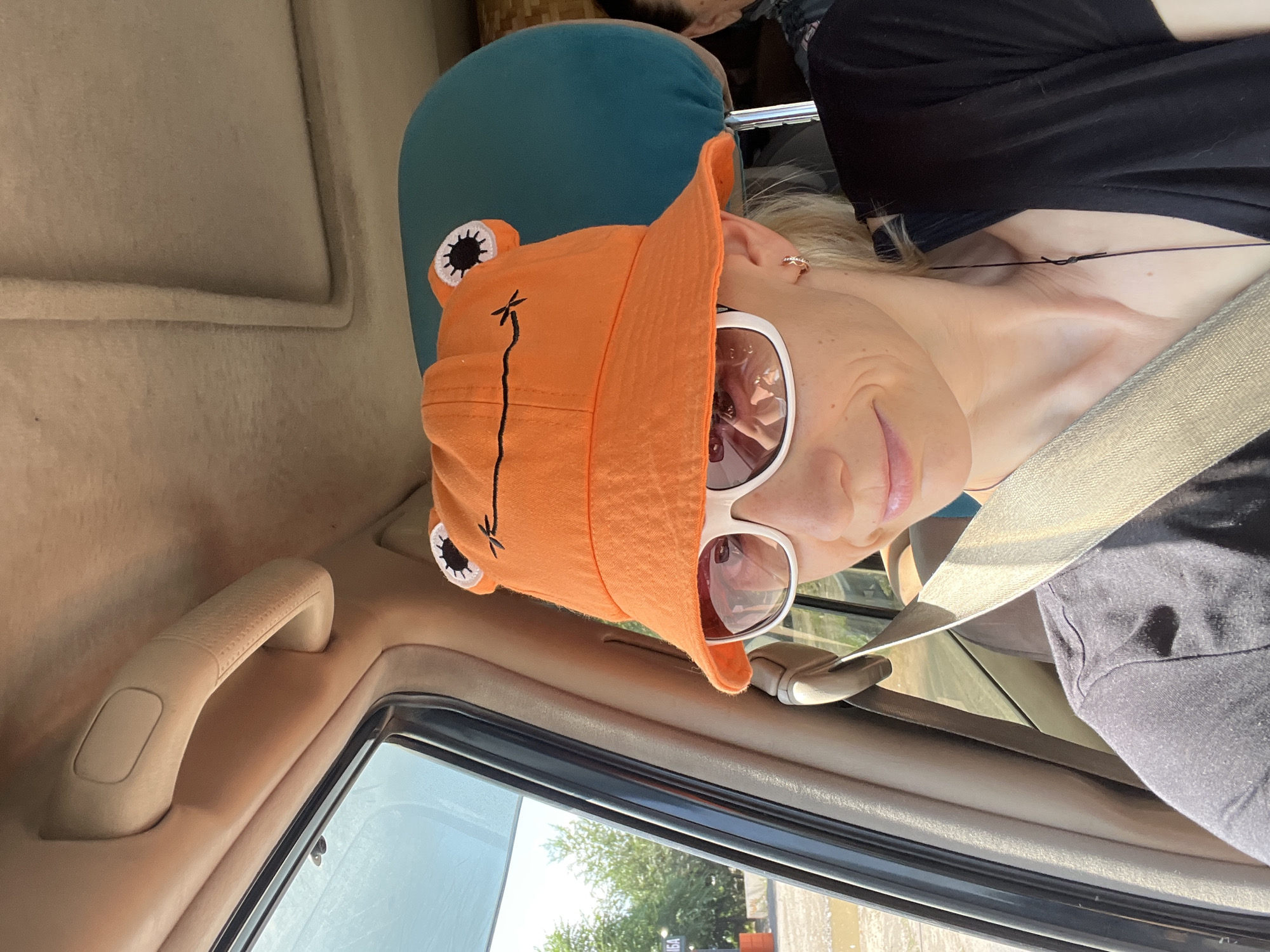 A woman wearing an orange bucket hat with frog eyes and a black t-shirt is sitting in the passenger seat of a vehicle. She is wearing white-rimmed sunglasses and a seatbelt. The interior of the car is visible, including the headrest of the seat in front of her and the car's ceiling. Outside the window, trees and a sign can be seen. The image appears to have been taken in Almaty, Kazakhstan, although no specific landmarks are identifiable.