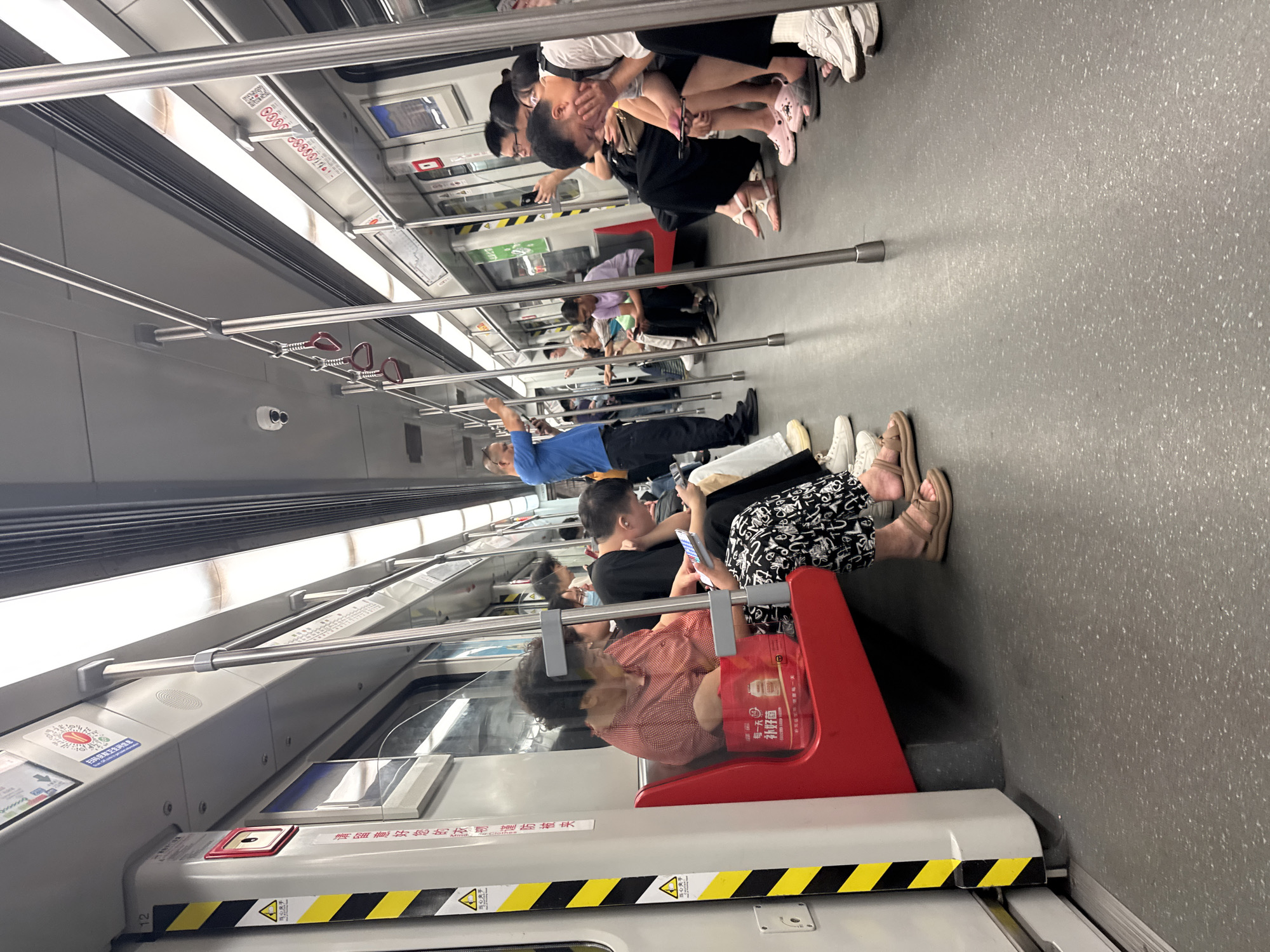 This image depicts the interior of a subway train carriage in Haizhu District, China. The carriage is moderately crowded with passengers seated on both sides of the aisle and standing near the doors. The seats are red and the floor is gray. Numerous metal poles are present throughout the carriage, serving as handrails for standing passengers. Several individuals are seen looking at their mobile phones. A man in a blue shirt stands near the door, holding his phone. The lighting appears to be artificial, coming from overhead fixtures. Yellow and black striped safety markings are visible on the side of the train car. Posters and advertisements are affixed to the interior walls.