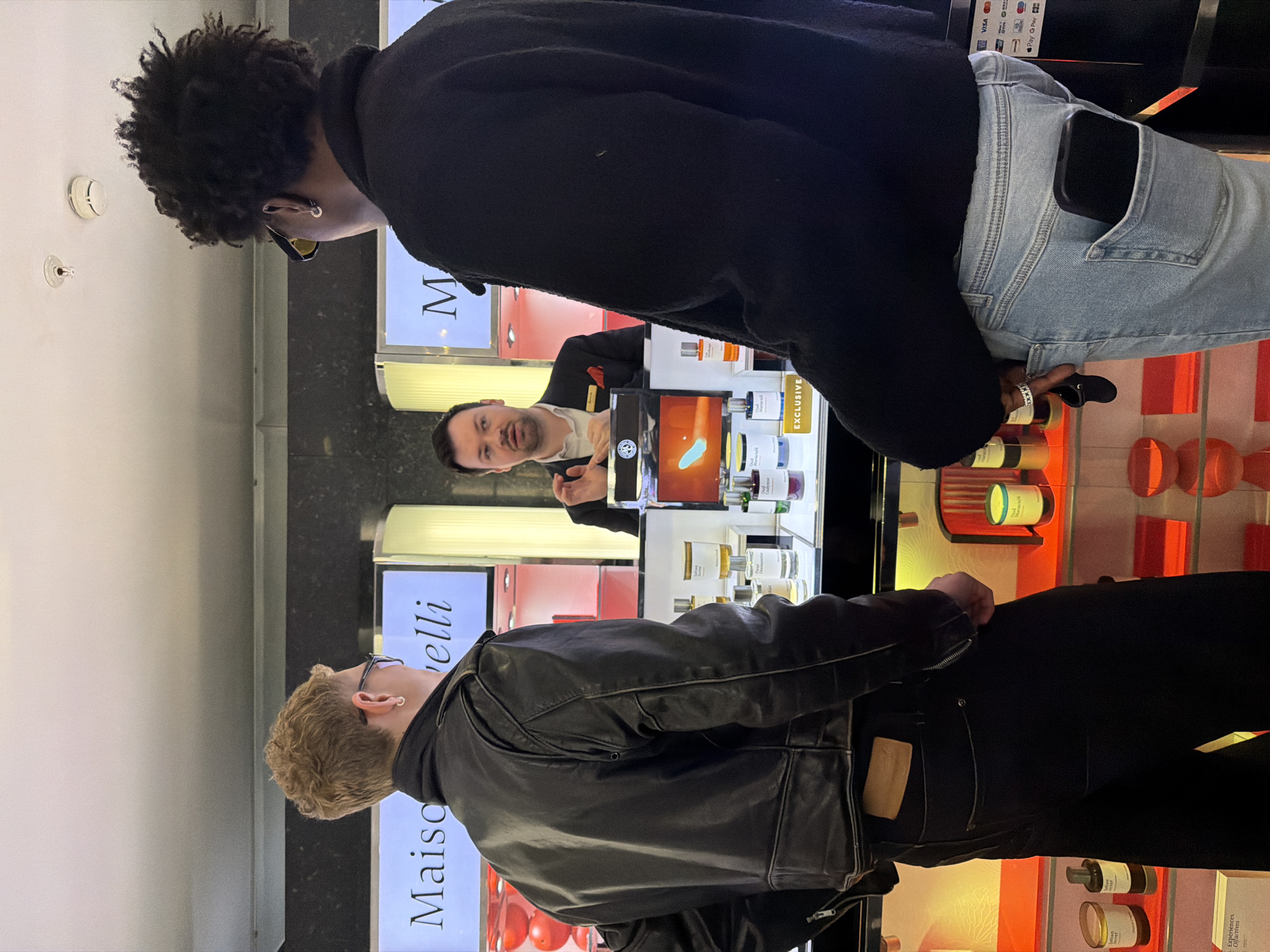 The image displays an indoor retail environment, specifically a perfumery counter, likely within a luxury department store in London, United Kingdom. Three adult males are present, engaged in a sales interaction.
At the center, behind a brightly illuminated product display counter, stands a male salesperson. He has short dark hair, a clean-shaven face, and is wearing a dark suit jacket over a white collared shirt, accessorized with a red pocket square. His hands are slightly raised and open, indicating he is speaking or explaining something to the customers.
Two male customers stand with their backs mostly to the camera, facing the salesperson. The customer on the left has short, wavy blonde hair, wears clear-framed glasses, a silver hoop earring in his left ear, a black leather jacket, and dark trousers with a light brown label visible on the rear waistband. The customer on the right has dark, curly hair, wears yellow-tinted sunglasses, a gold hoop earring in his right ear, a dark long-sleeved top, and light blue denim jeans, with a black smartphone visible in his right rear pocket.
The counter features numerous perfume bottles, candles, and other scented products, with prominent backlighting. Large, illuminated signs displaying "Maison Crivelli" are visible in the background behind the salesperson. A smaller illuminated sign on the counter reads "EXCLUSIVE". On the far right, a payment terminal is partially visible, showing logos for Visa, Mastercard, American Express, Apple Pay, and G Pay. The surrounding environment features dark walls with integrated lighting and reflective surfaces, while the white ceiling shows two circular ceiling-mounted devices, possibly a smoke detector and a sprinkler head. Red spherical decorative items are visible on shelves to the left.