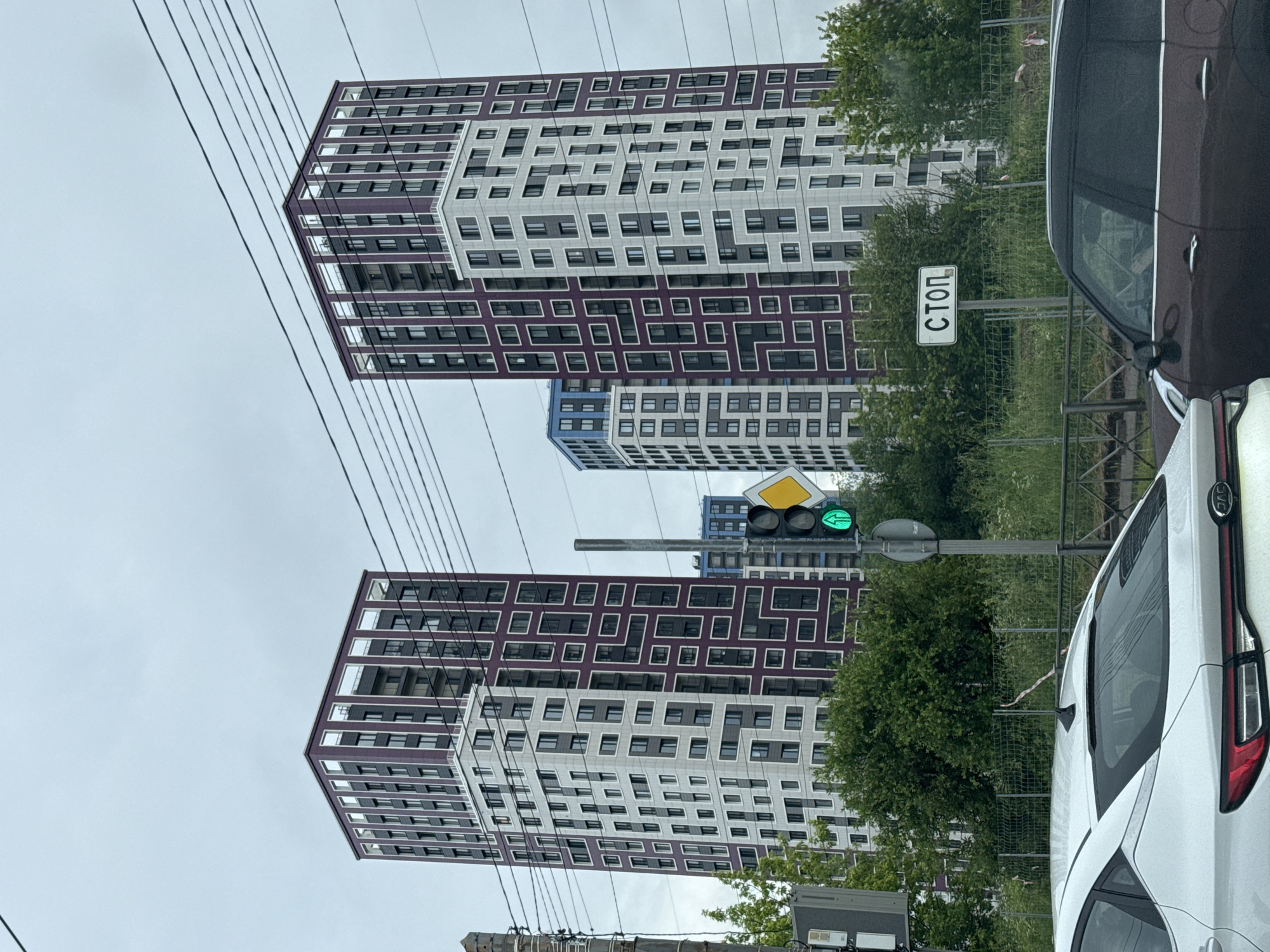 The image shows a street scene in Saint Petersburg, Russia, with two tall modern residential buildings as the central focus. The buildings have a distinctive facade design with alternating white and dark purple rectangular patterns surrounding numerous windows. A third, shorter building with a blue upper section is visible behind the main structures. In the foreground, the rear of a white car and a dark brown car are visible. To the right, a traffic light is illuminated with a green signal, and a diamond-shaped road sign with a yellow border is present. A white rectangular sign with black Cyrillic text reading "СТОП" (STOP) is also visible on a pole. The sky is overcast, and power lines are strung across the upper portion of the image. Lush green trees and vegetation border the street and fence in the middle ground. The scene appears to be captured from inside another vehicle, looking out.