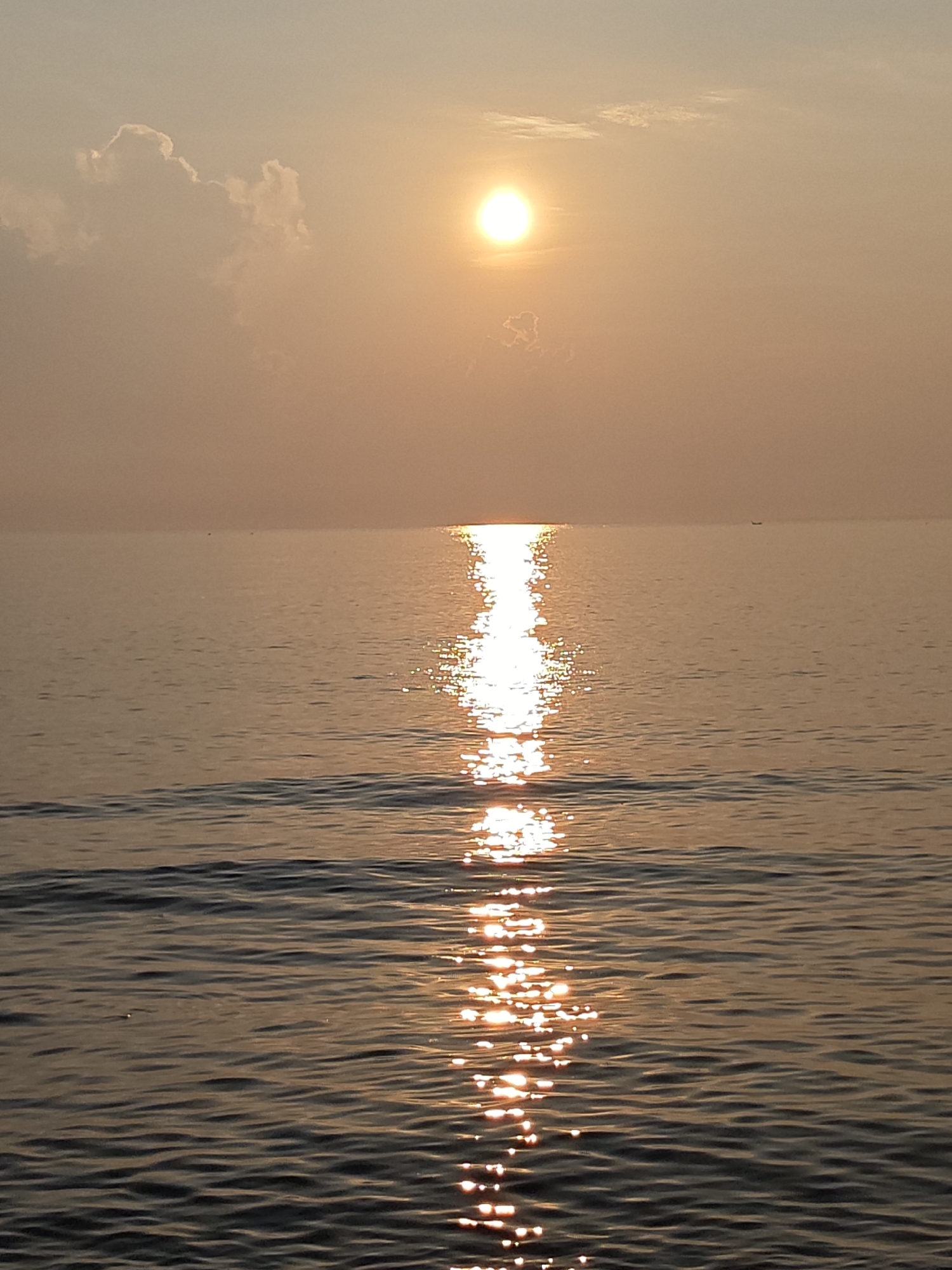 The image depicts a sunset over water, likely a sea or large lake. The sun is positioned high in the sky, with its bright reflection stretching across the water towards the viewer. The sky has a hazy, golden hue, and there are a few scattered clouds near the sun. The water's surface is rippled, and the reflection of the sun appears as a shimmering path of light. In the distance, there is a hint of a small boat on the horizon. It is difficult to confirm the exact location as Lhokseumawe, Indonesia without landmarks or more specific environmental clues.