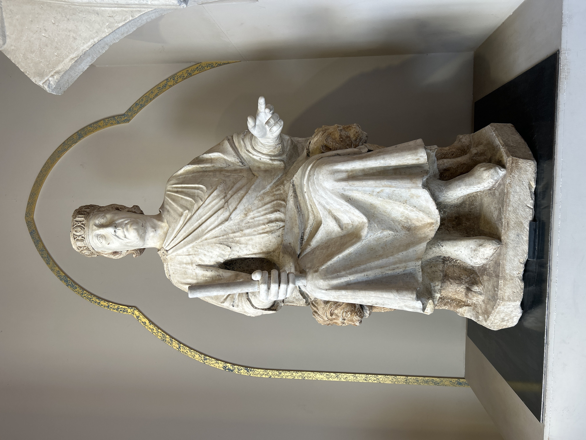 A large, light-colored stone statue, identified as Pope Boniface VIII by Arnolfo di Cambio, is centrally positioned against a plain, light beige wall. The crowned male figure is seated, draped in flowing robes that obscure his lower body, holding a cylindrical object, likely a scepter or scroll, in his left hand. His right hand is extended with the index finger pointed forward. The figure sits upon a throne adorned with two visible lion heads supporting the armrests. The statue's rough-hewn octagonal base rests on a dark rectangular mat, which is placed on a wider, light-colored plinth. An ornate golden arch, featuring patterned blue and black details, frames the upper part of the statue, originating from the wall behind it. A segment of a light-colored architectural structure is partially visible on the far right. This artwork is located within the Palazzo dei Conservatori, part of the Capitoline Museums in Rome, Italy.