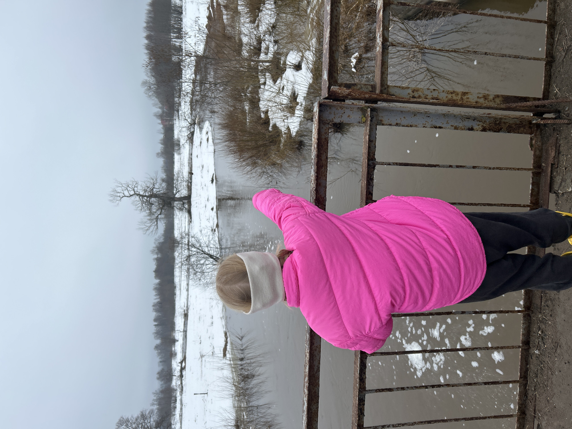 A child in a bright pink winter coat, black pants, and a white headband is standing on a metal grate structure, possibly a bridge or platform, overlooking a body of water. The water is surrounded by snowy terrain and bare trees, suggesting a cold or winter season. The sky is overcast. The scene appears to be in Уральское, Russia.