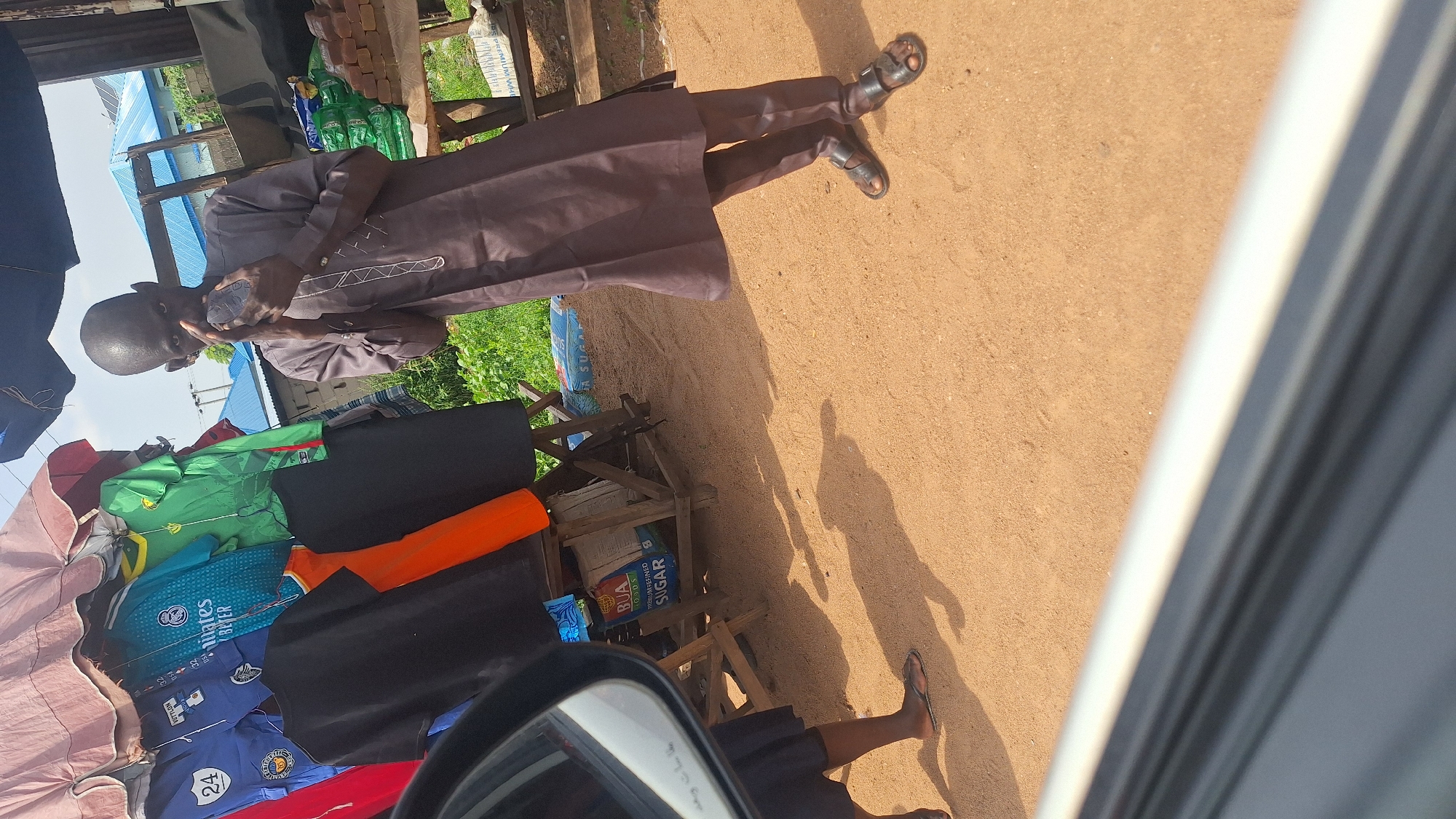 In Jalingo, Nigeria, a man stands on a sandy path, wearing a brown agbada and dark sandals. He is holding what appears to be a phone to his face. Behind him, a stall displays various sports jerseys, including blue, green, and yellow ones. To the right of the stall, there are sacks of sugar and a pile of bricks. The ground is covered in fine sand, and shadows are cast by the stall and its contents. The sky is visible in the background, with some greenery and buildings.