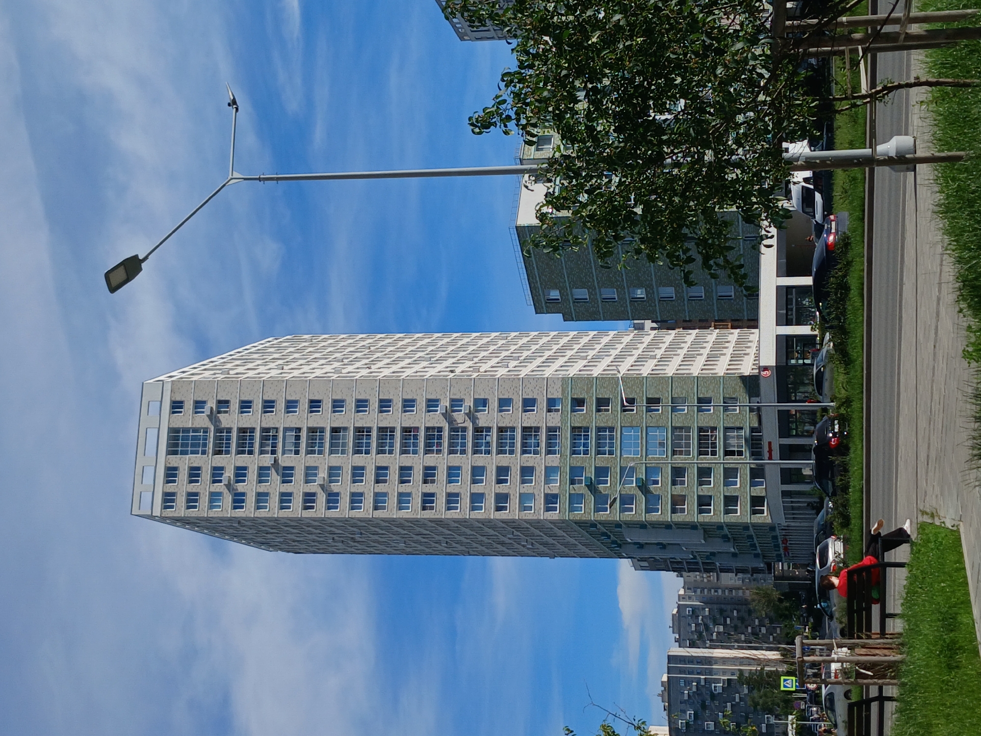 In Yekaterinburg, Russia, a tall, modern apartment building dominates the frame, its light gray facade punctuated by numerous windows. To the right, a shorter, similarly styled building features a green tiled section. A solitary individual is seated on a park bench in the foreground, facing away from the camera, seemingly observing the street. Several cars are parked or driving on the road. A lamppost stands prominently on the right side of the image. The scene is set against a clear blue sky with scattered white clouds, under a bright daylight condition. Lush green grass and some trees add to the urban landscape.