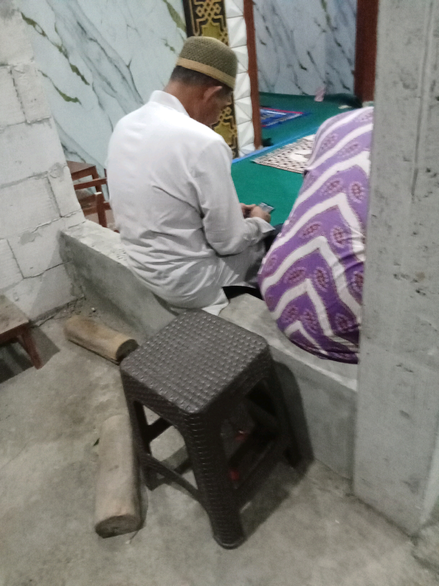 In the city of Keplaksari, Indonesia, a man wearing a white garment and a green cap sits facing away from the camera, looking at his phone. Beside him, a woman in a purple and white patterned garment is also seated. They are inside what appears to be a mosque, indicated by the prayer rugs visible in the background. In the foreground, a dark brown plastic stool sits on the concrete floor, with a wooden log lying nearby. The walls feature a marble-like pattern. The lighting is dim, suggesting it might be evening.