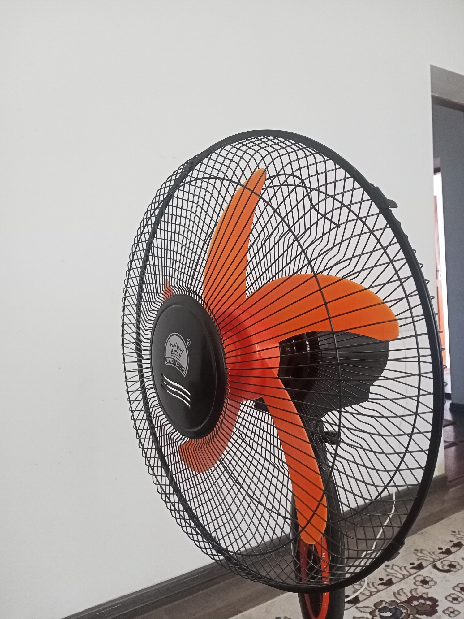 The image shows a close-up view of an electric fan with orange blades and a black grille. The fan is positioned in front of a plain white wall, with a portion of a doorway and a rug visible in the background. The fan has a circular logo on the front grille which reads "CHANGLI CROWN". There are no people or animals present in the image. The scene appears to be an indoor setting, likely a room in a residential or commercial building in the city of Sokuluk, Kyrgyzstan.