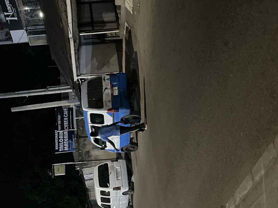 A street scene at night features two white and blue vans parked near a building. One van is occupied by a person, seen through the open door, seemingly entering or exiting. A sign for "Honeysmart Tailoring Material Cyber Cafe" is visible. The road surface is dark, with shadows obscuring details. The perspective is from a high angle, rotated counterclockwise.