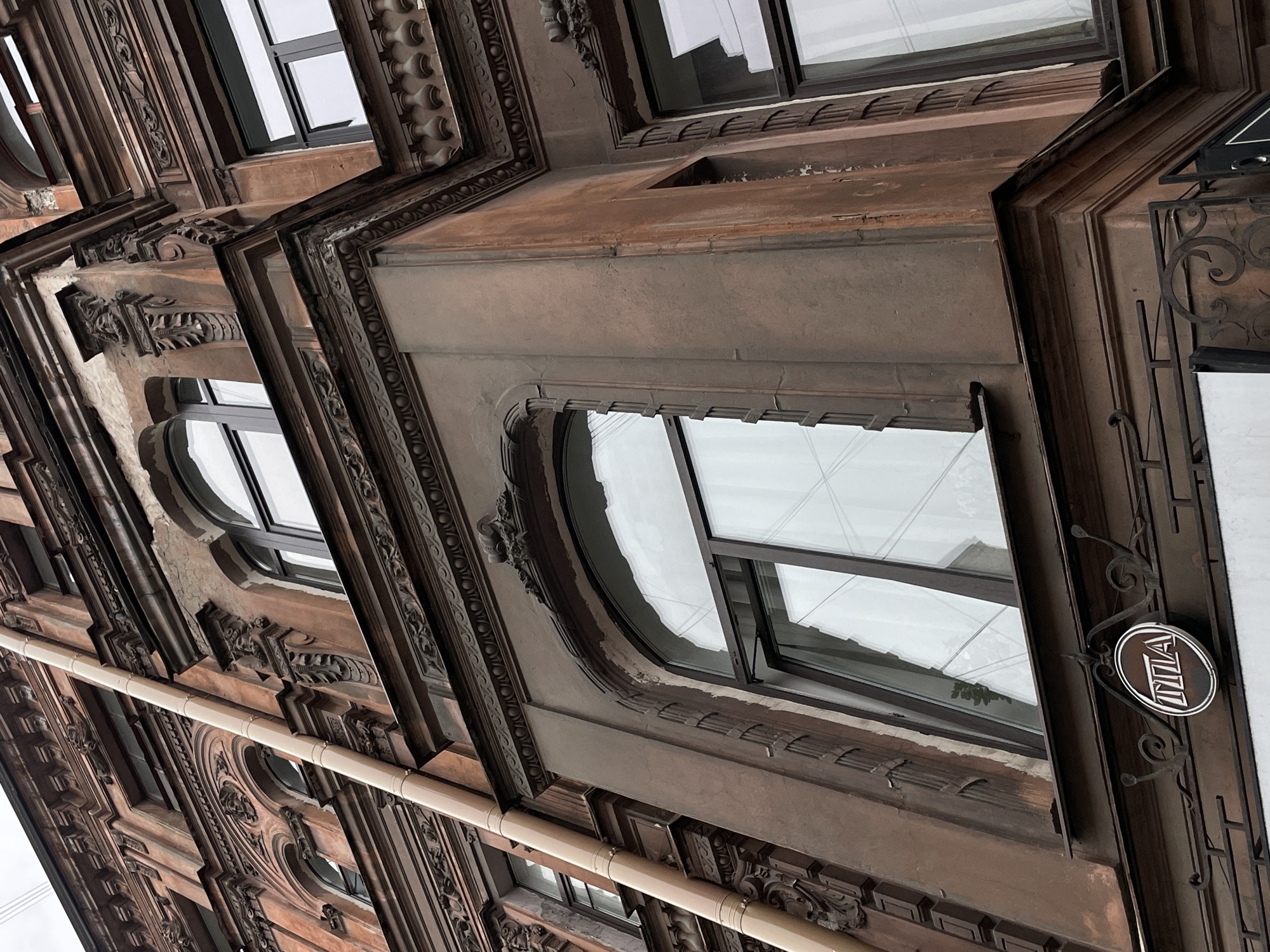 The image shows the facade of a building, likely in Saint Petersburg, Russia, based on the architectural style and the sign reading "ПЛА" (PLA), which could refer to a local organization. The building features ornate detailing around the windows, including moldings, carvings, and decorative elements. The facade appears to be made of a brownish-gray material. The windows have a modern, dark-framed design. A light-colored pipe runs horizontally across the building's exterior. The photograph is taken from a low angle, looking upwards. The overall impression is that of a historic building with modern updates.