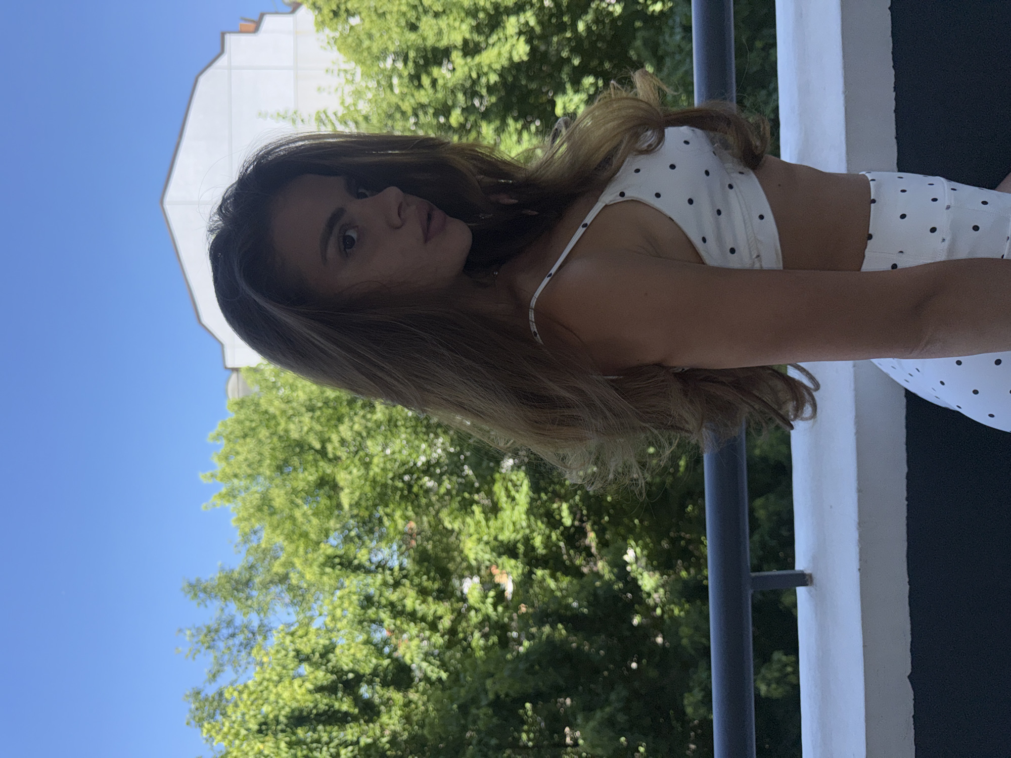 A young woman with long, wavy brown hair stands on a balcony, looking to her right. She is wearing a white bikini top with black polka dots and a matching white skirt. In the background, lush green trees and a white building are visible under a clear blue sky. The scene appears to be a warm, sunny day. This balcony could be located anywhere in Plovdiv, Bulgaria, with similar architecture and greenery.