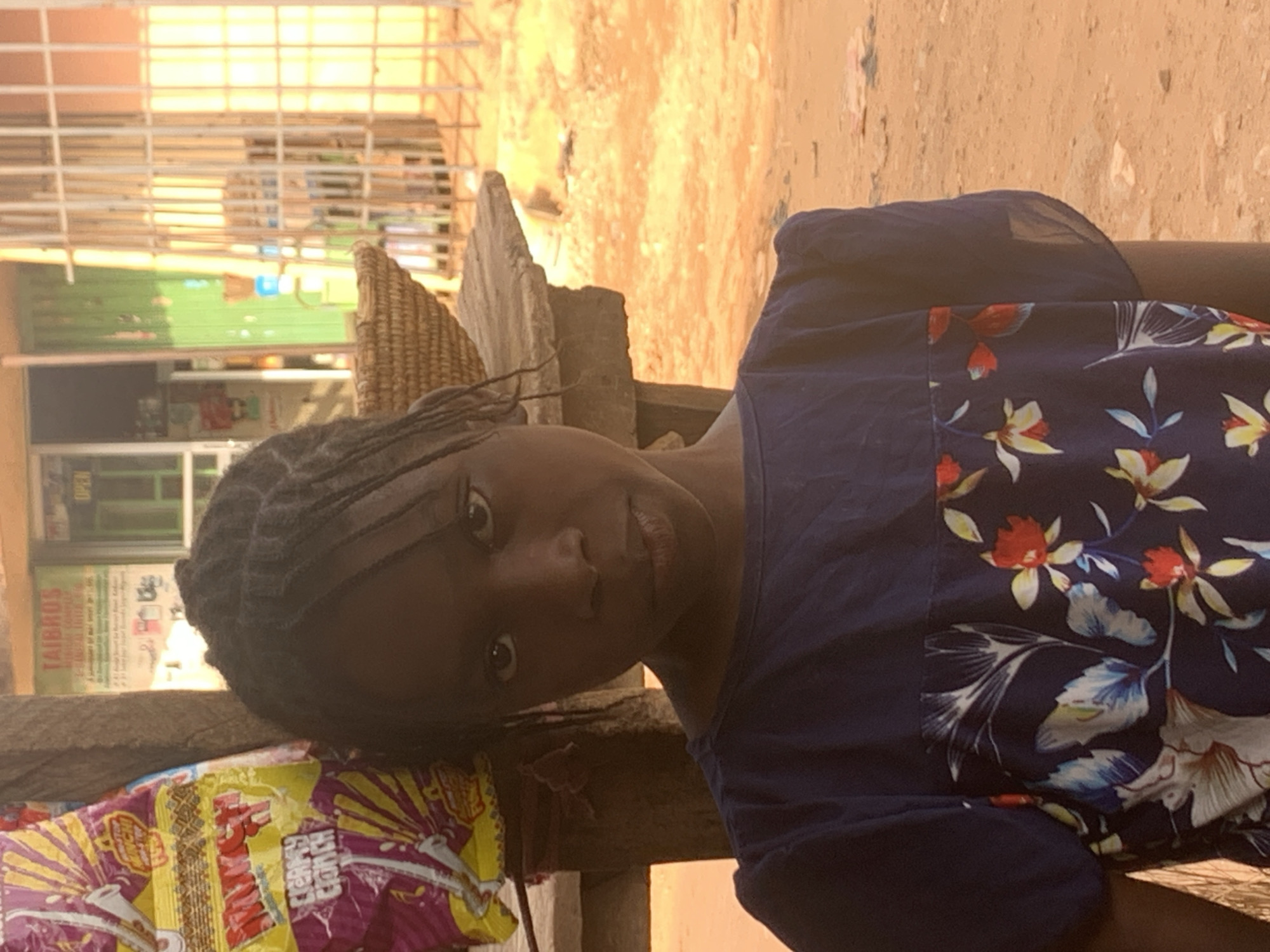 A girl with braided hair is seated on a wooden structure, possibly a bench or stool, in what appears to be an outdoor setting. The location is suspected to be in Sabon Tasha, Nigeria. She wears a blue shirt with floral patterns. To the left, there is a colorful bag of snacks, possibly "Creamy Crunch". In the background, there is a building with a green facade, and a storefront with signs labeled "TAI BRO". The scene is captured in daylight, with the light source positioned in front of the subject, causing some glare and bright highlights in the image. The surrounding environment appears to be a street or public space, with evidence of other structures and activity in the background.