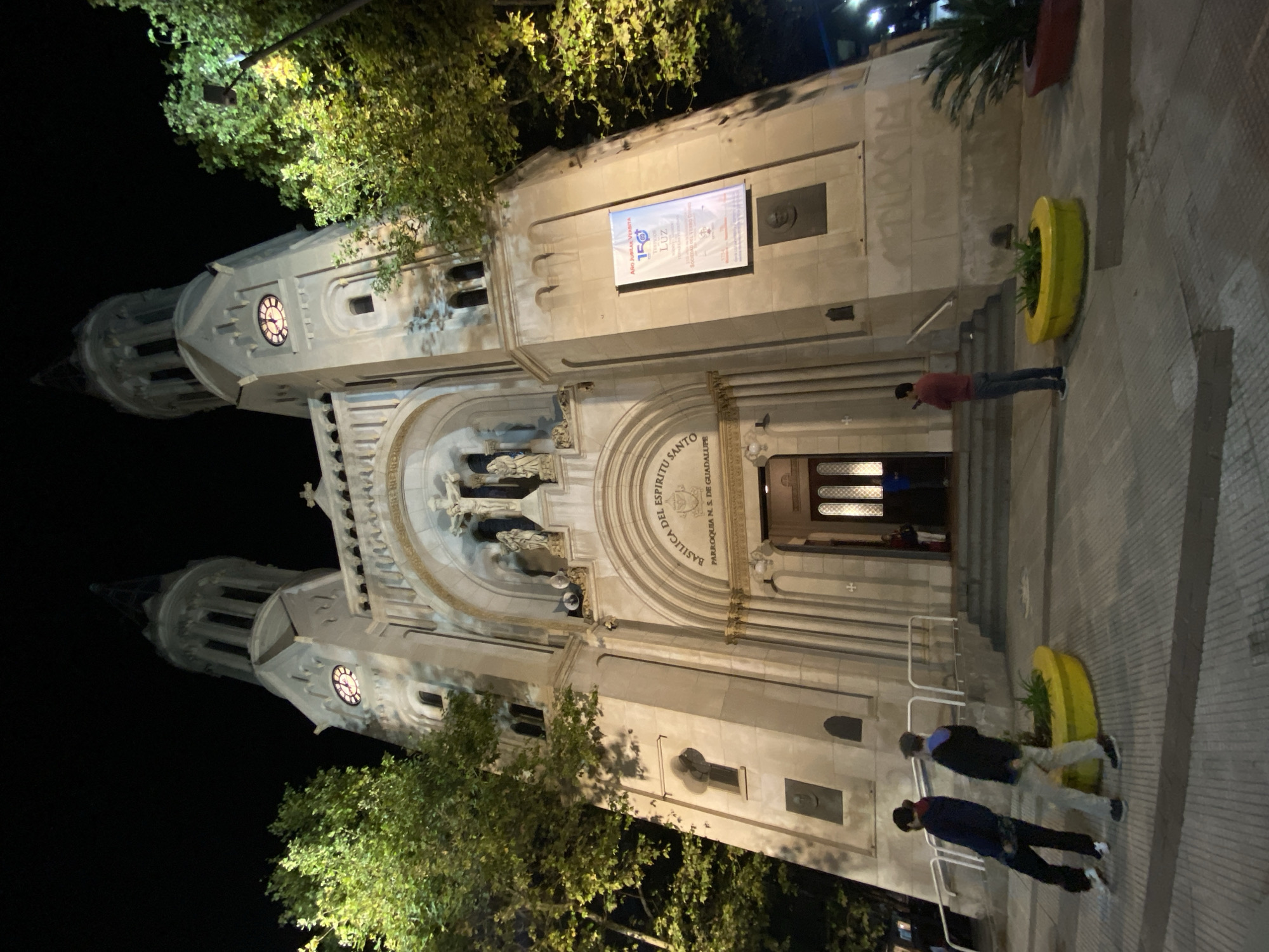 The image captures the Basilica del Espiritu Santo in Buenos Aires, Argentina, at night. The building is illuminated, showcasing its architectural details, including two towers with clocks and statues above the entrance. A banner celebrating an anniversary is visible on the building's facade. Three people are walking in front of the basilica, and one person is standing near the entrance. Yellow planters flank the steps leading to the entrance. Trees frame the basilica on both sides, their leaves reflecting the artificial light. The night sky is dark.