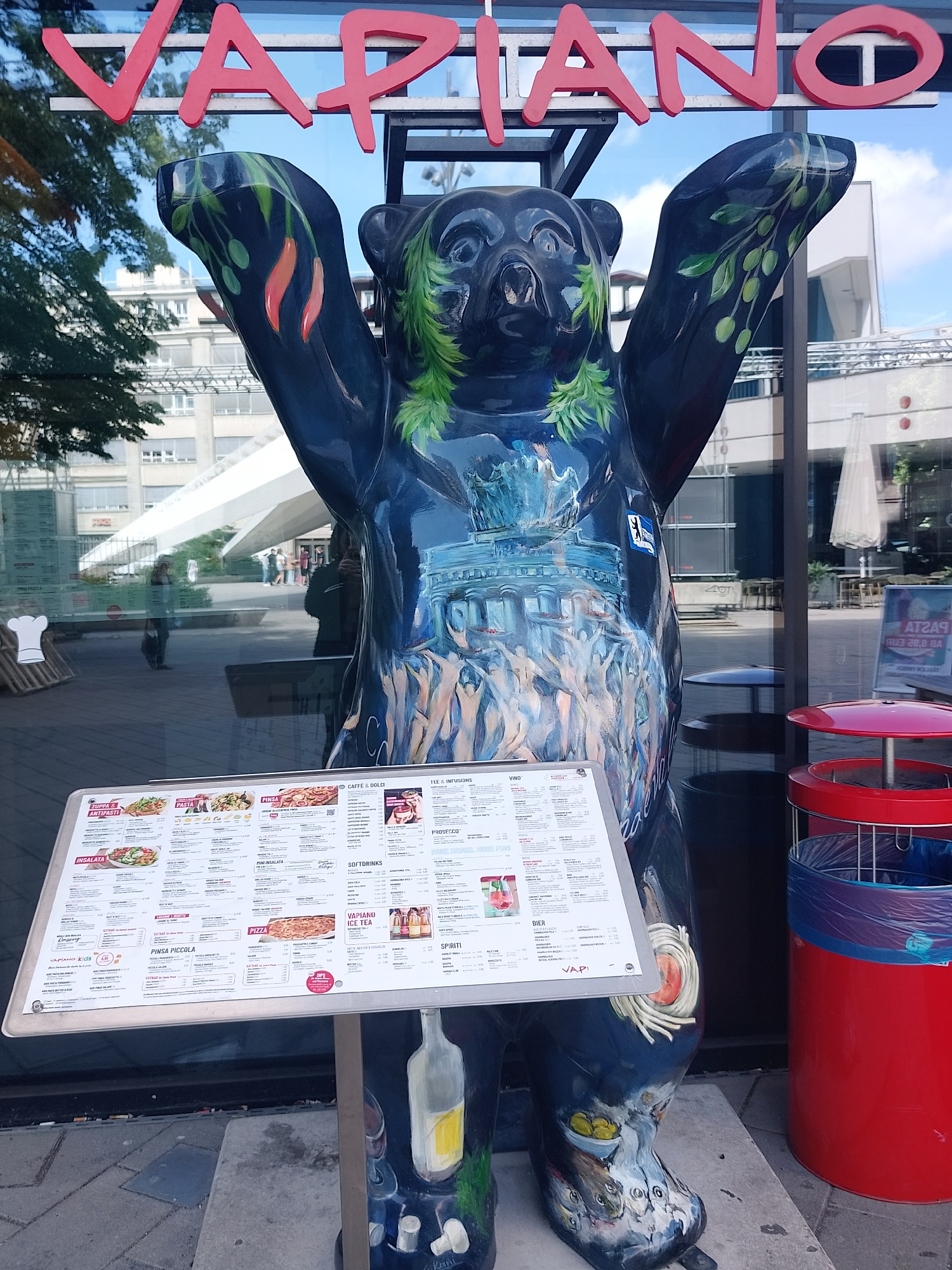 The image captures a Vapiano restaurant in an unidentified city. In front of the restaurant, a painted bear statue stands, holding a menu board. The bear is decorated with images of a fountain and figures, and has greenery painted around its neck and limbs. The restaurant's sign, "VAPIANO," is visible above the bear. A red trash can is placed to the right of the statue. Through the restaurant's glass façade, hints of a modern architectural structure can be seen in the background, suggesting an urban environment.