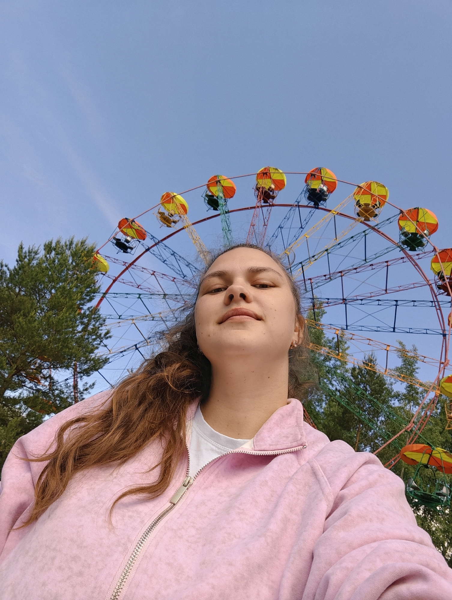 A medium shot captures a young woman in front of a Ferris wheel. The Ferris wheel, painted in vibrant red, orange, yellow and blue, is partially obscured by green trees and dominates the background. The woman, positioned in the foreground, has long brown curly hair, and wears a pink zippered jacket over a white t-shirt. The sky is a clear light blue. The location is Chaikovsky, Russia.