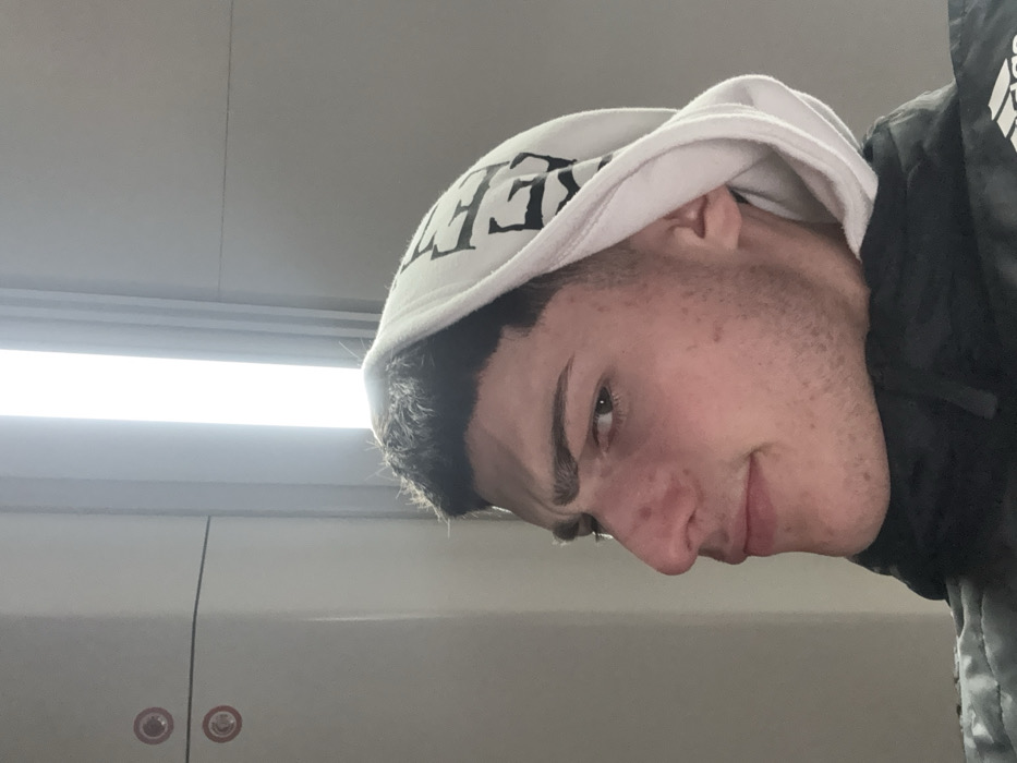 A young man with short dark hair and fair skin looks down at the camera. He has a slight smile. He is wearing a white hoodie with black lettering. Part of a dark jacket with white detailing is visible on the right. The background is a light grey surface and a light fixture. Two red circles with white smiley faces appear in the lower left corner.