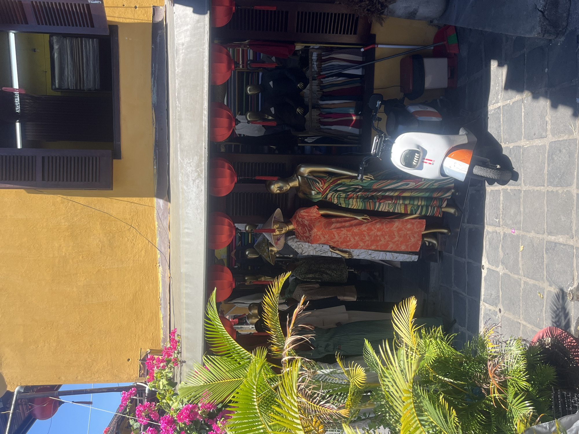 The image presents a vibrant street scene in Hội An, Vietnam, depicting the facade of a clothing shop under bright daylight. The building features a distinctive yellow-painted wall with an open window on the upper left, revealing curtains and dark wooden shutters. Below, the shop's entrance is decorated with several red lanterns suspended from an awning.
Within the open storefront, multiple mannequins are displayed, showcasing a variety of colorful garments, including patterned dresses that appear to be traditional Vietnamese styles. Some mannequins wear conical hats (nón lá). Behind them, shelves and racks are filled with neatly folded and hung clothing items.
To the right of the shop entrance, a white and orange scooter is parked on the gray tiled pavement. The text "M16" is visible on its side. Nearby, cleaning tools, specifically a broom and a dustpan, lean against the wall. Lush green plants with palm-like fronds and clusters of vibrant pink bougainvillea-like flowers are visible in the foreground, adding to the tropical atmosphere. Strong sunlight casts distinct shadows on the ground and surfaces, indicating a sunny day, likely around midday.
