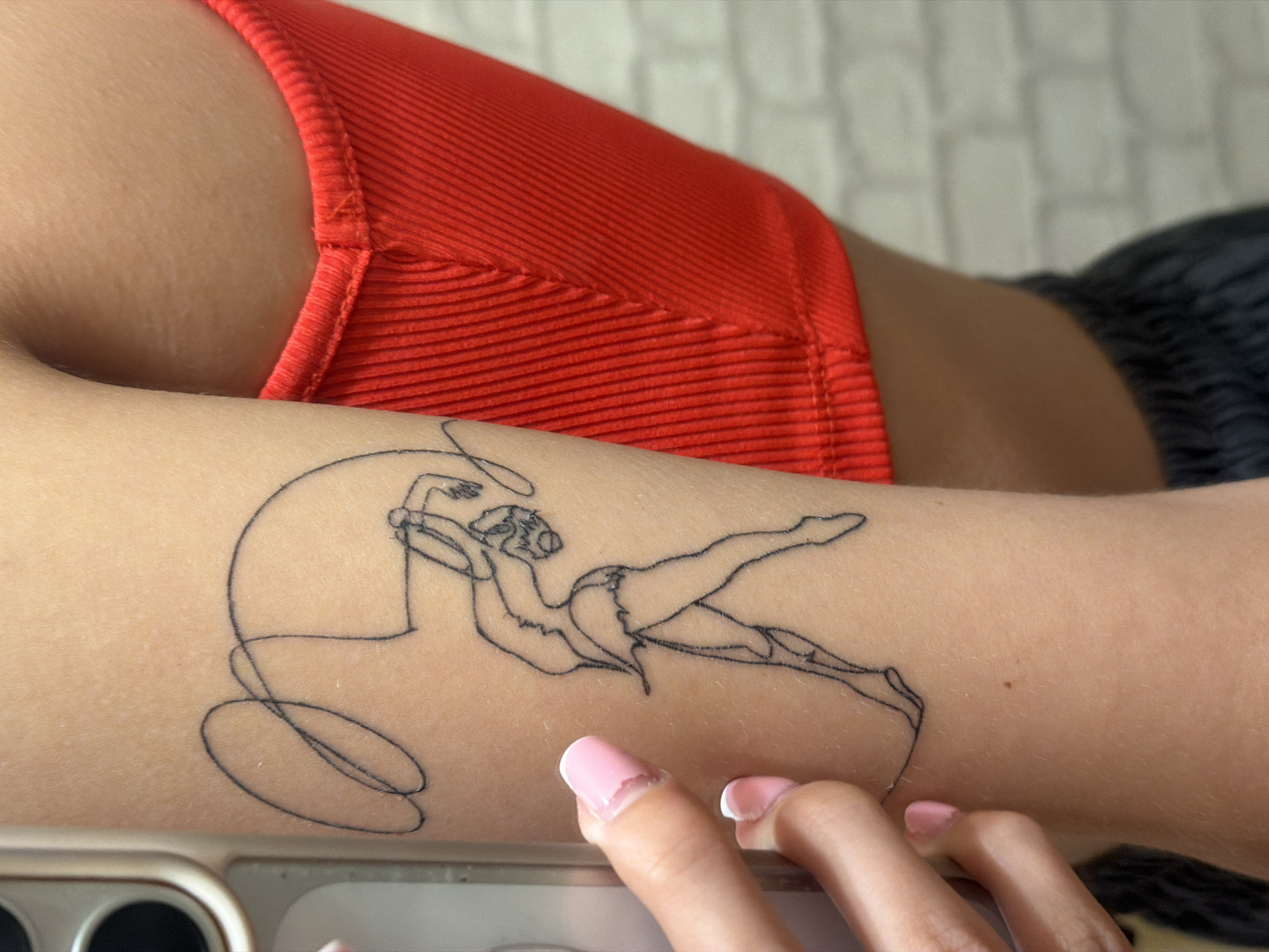 The image displays a close-up view of a person's arm, specifically the upper arm, where a thin-lined black tattoo of a gymnast with a ribbon is visible. The tattoo is depicted in a minimalist style, outlining the gymnast's body and the flowing ribbon. The individual is wearing a bright red ribbed tank top, and a portion of dark gray athletic shorts can be seen at the bottom of the frame. The skin tone is fair, and one finger with a light pink nail polish is touching the tattoo. The background is out of focus, showing a hint of a white brick wall. The overall lighting suggests an indoor setting. The location is not specified in the image or its context.
