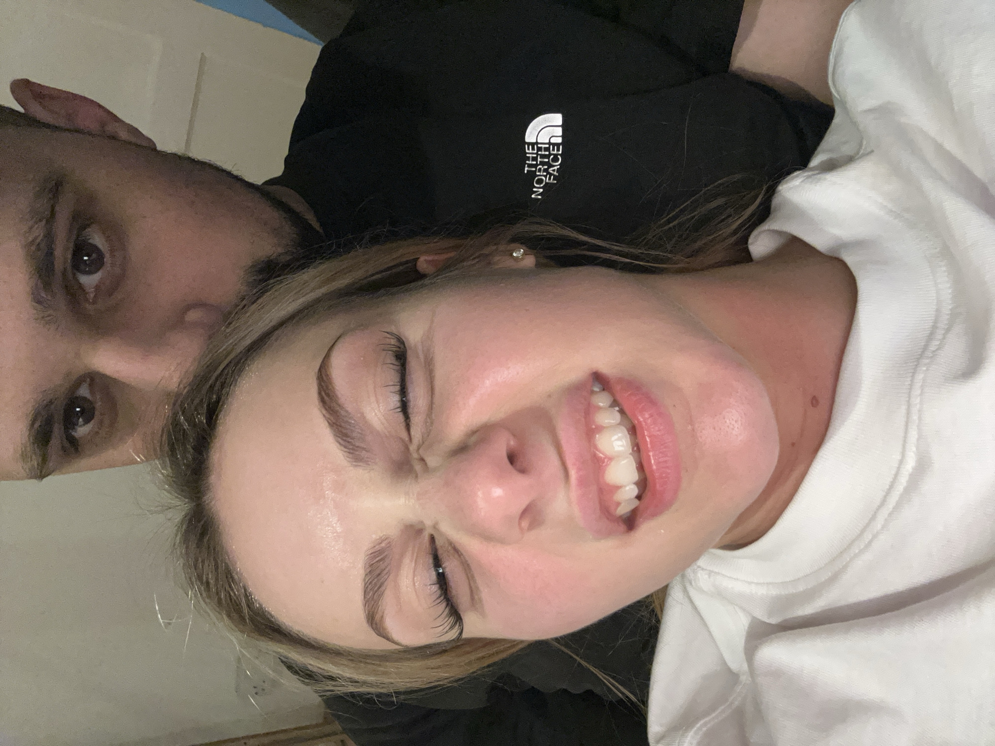 Two people are visible in the image. One person, seemingly male, is in the background, partially visible with dark hair and a black shirt with the logo "The North Face". The other person, seemingly female, is in the foreground wearing a white shirt. She is smiling with her eyes closed, displaying her teeth. She has fair skin, with professionally done eyelashes. The location is in the city of Unknown, Unknown.