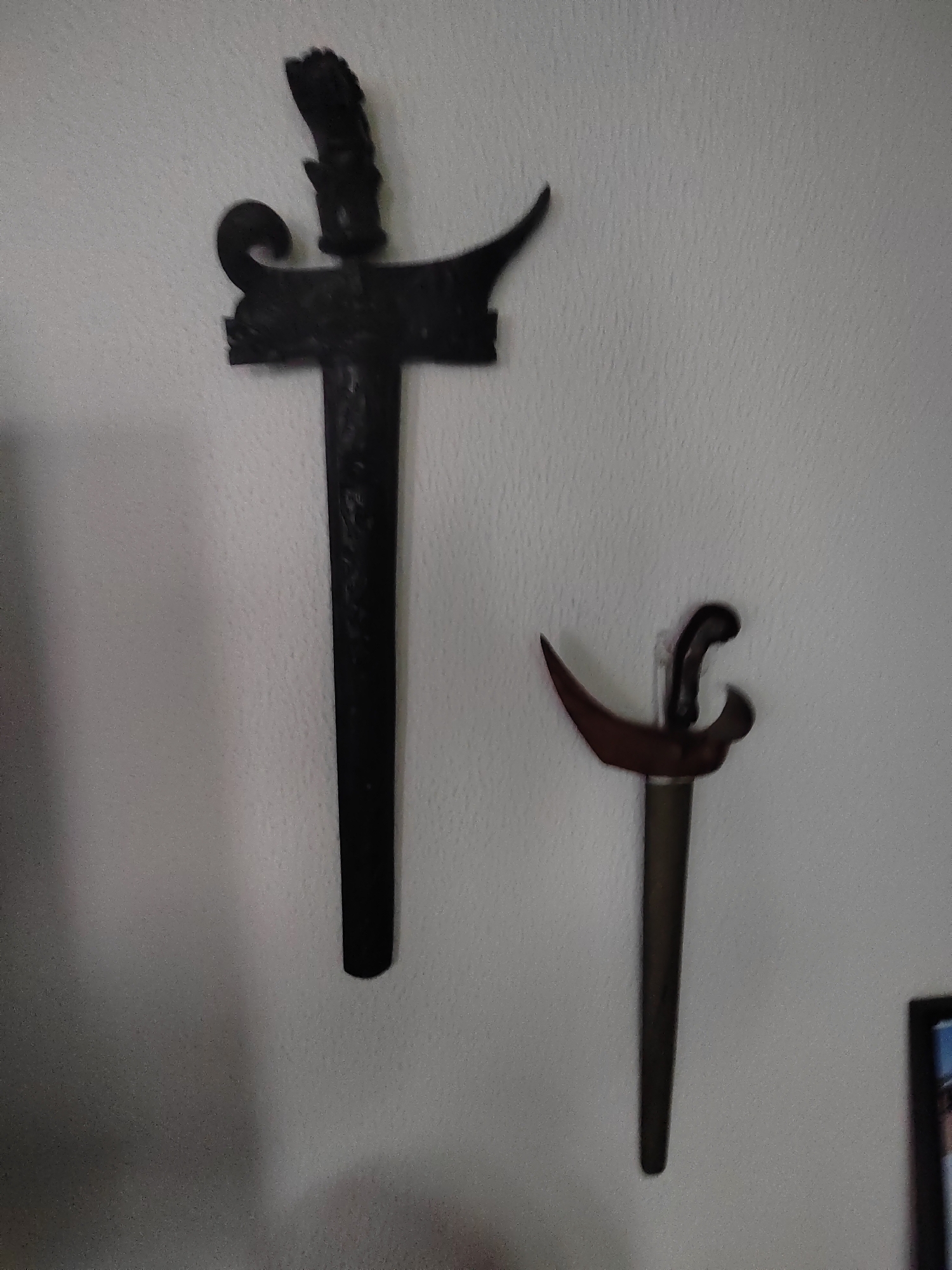 Two daggers are mounted on a textured, off-white wall. The dagger on the left is larger and appears to be made of dark metal. It has a wavy blade and an ornate hilt. The dagger on the right is smaller and also made of dark metal, with a curved blade and a wooden hilt. Both daggers are positioned vertically. The environment is indoor, with the wall providing the primary background. The lighting appears to be ambient and slightly diffused. There is a hint of a framed picture on the lower right of the image. The location is identified as Porto Recanati, Italy.