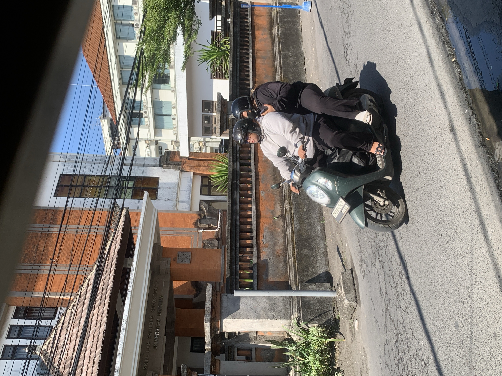 Two individuals wearing helmets are riding on a green scooter. The person in the front is wearing a light gray sweatshirt and black pants. The person in the back is wearing a black sweatshirt and black pants, and has their face partially obscured by a black mask. They are on a paved road in front of a building with an orange tiled roof and a white facade. The building has visible signage that reads "SKILL LAB FAKULTAS KEDOKTERAN UNIVERSITAS UDAYANA". The environment suggests an urban setting, likely in Denpasar, Indonesia, given the building's signage. The road surface is cracked and worn, with a shadow cast across it. To the right of the scooter, a blue pole with a white cap is visible. Lush green vegetation can be seen along the wall behind the scooter.