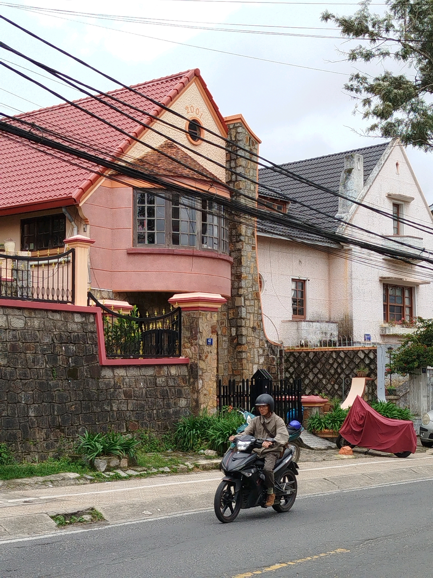 A person wearing a helmet rides a black motorcycle down a paved street in Da Lat, Vietnam. The motorcycle is in the foreground, with the rider facing towards the camera. Behind the motorcycle, there are two houses. The house on the left has a pink facade and a red tiled roof. The house on the right has a white facade and a dark gray tiled roof. Both houses have multiple windows and architectural details. Overhead, numerous electrical wires crisscross the sky. The street is lined with a stone wall and fences, with some greenery and plants visible. The sky is overcast. A red tarp covers another motorcycle parked on the side of the road.