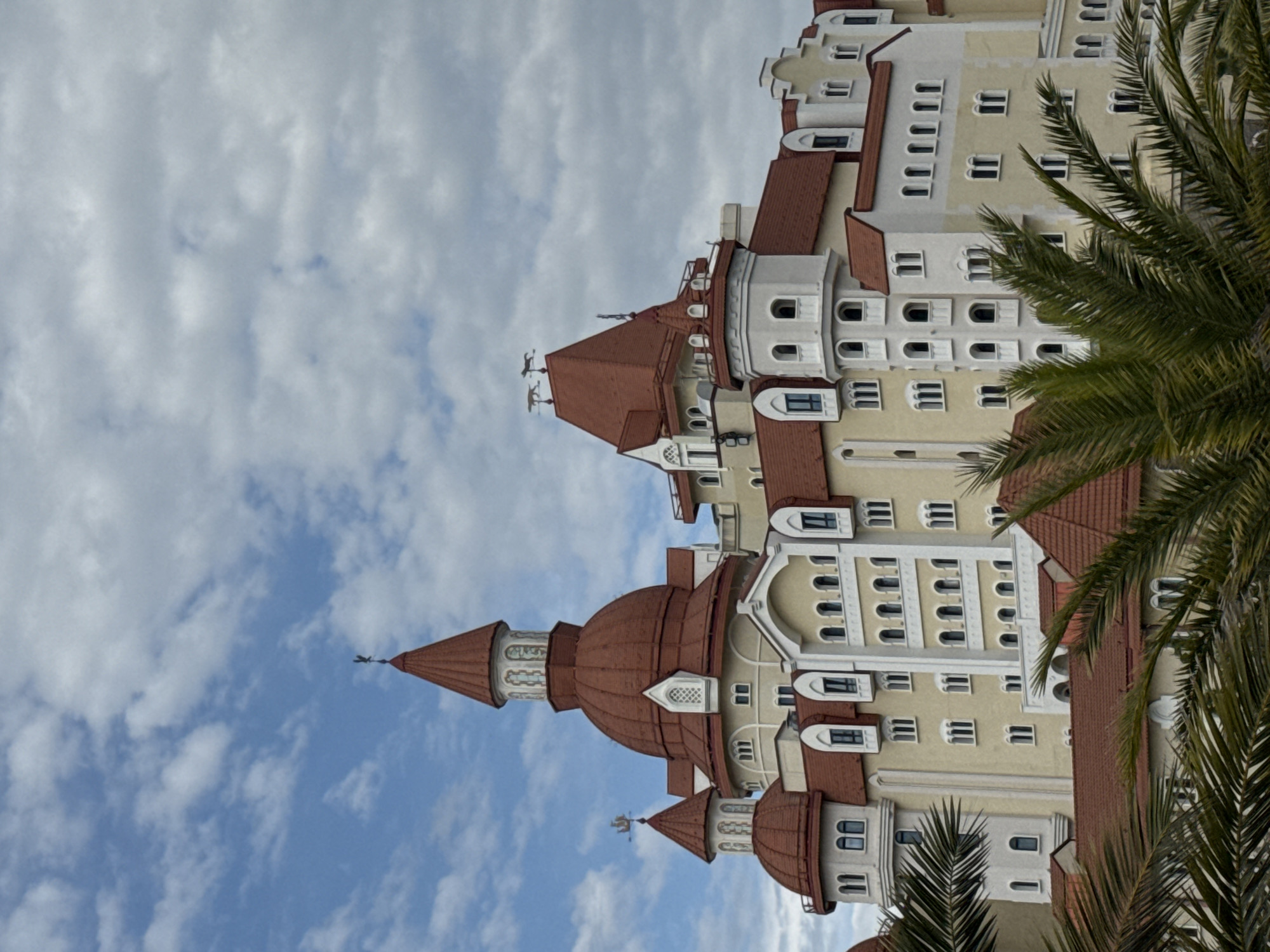 The image shows a building with multiple towers and red-tiled roofs, partially obscured by palm trees. The building has a light yellow facade with white architectural details and numerous windows. The sky is visible in the background, with a mix of blue and white clouds. The location is identified as the city of Sirius, Russia.