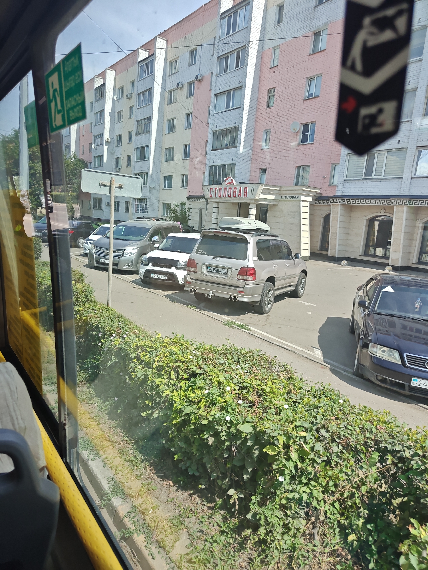 The image, taken from inside a vehicle, captures a street scene in Petropavl, Kazakhstan, on a sunny day. The foreground is dominated by a view through a window, with the yellow interior of the vehicle visible on the left. Beyond the window, a row of parked cars lines the street in front of a multi-story residential building. The building exhibits a pastel color scheme, with sections painted in light pink, yellow, and white. Several windows, some with air conditioning units, are visible on the building's facade. A green emergency exit sign with pictograms and text is affixed to the vehicle's interior, partially obscuring the view.
Several vehicles are parked along the street. Closest to the building is a silver SUV with a roof rack, followed by a white compact car and a gray minivan. Further down the street, a dark blue sedan is parked. The vehicles have license plates, some of which are partially legible, appearing to be from Kazakhstan. A dense green hedge with small white flowers runs along the sidewalk in front of the parked cars. In the background, trees and the sky are visible. A black banner with a white emblem is hanging from an unseen structure, adding a decorative element to the scene. The overall impression is of a typical urban street in a residential area.