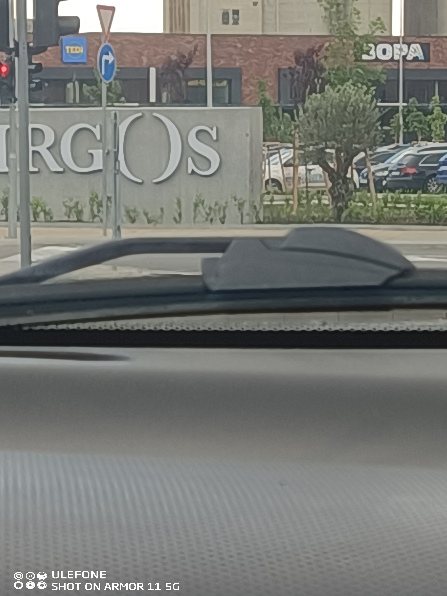 The image depicts a daytime urban scene viewed from the interior of a vehicle, looking through the windshield. In the immediate foreground, a grey, textured dashboard is visible, along with a black windshield wiper arm positioned horizontally and a dark grey housing unit, likely for a sensor, attached to the windshield.
In the midground, a street environment is visible. On the left, a traffic light pole with a red signal is present. Adjacent to it, a second pole supports two road signs: an upper triangular warning sign with a red border and a white interior, and a lower circular mandatory sign with a blue background and a white right-pointing arrow. Behind the traffic light pole, a blue rectangular sign with "TEDI" in white capital letters is visible.
A prominent building forms the central background. Its lower section features a large grey wall displaying the word "ARGOS" in white, serif capital letters. Above this, the building has a reddish-brown brick facade with dark window frames. To the right of the "ARGOS" sign, another sign on the brick facade reads "3OPA" in white capital letters. Small green foliage is planted along the base of the "ARGOS" wall. To the right of the center, an olive tree with a slender trunk and dense green leaves stands.
Further in the background, to the right of the olive tree, a parking area contains several parked vehicles, including what appears to be a dark-colored SUV or wagon. The sky is bright and appears overcast, providing diffuse daylight. No individuals are visible in the scene. The location is currently unknown.