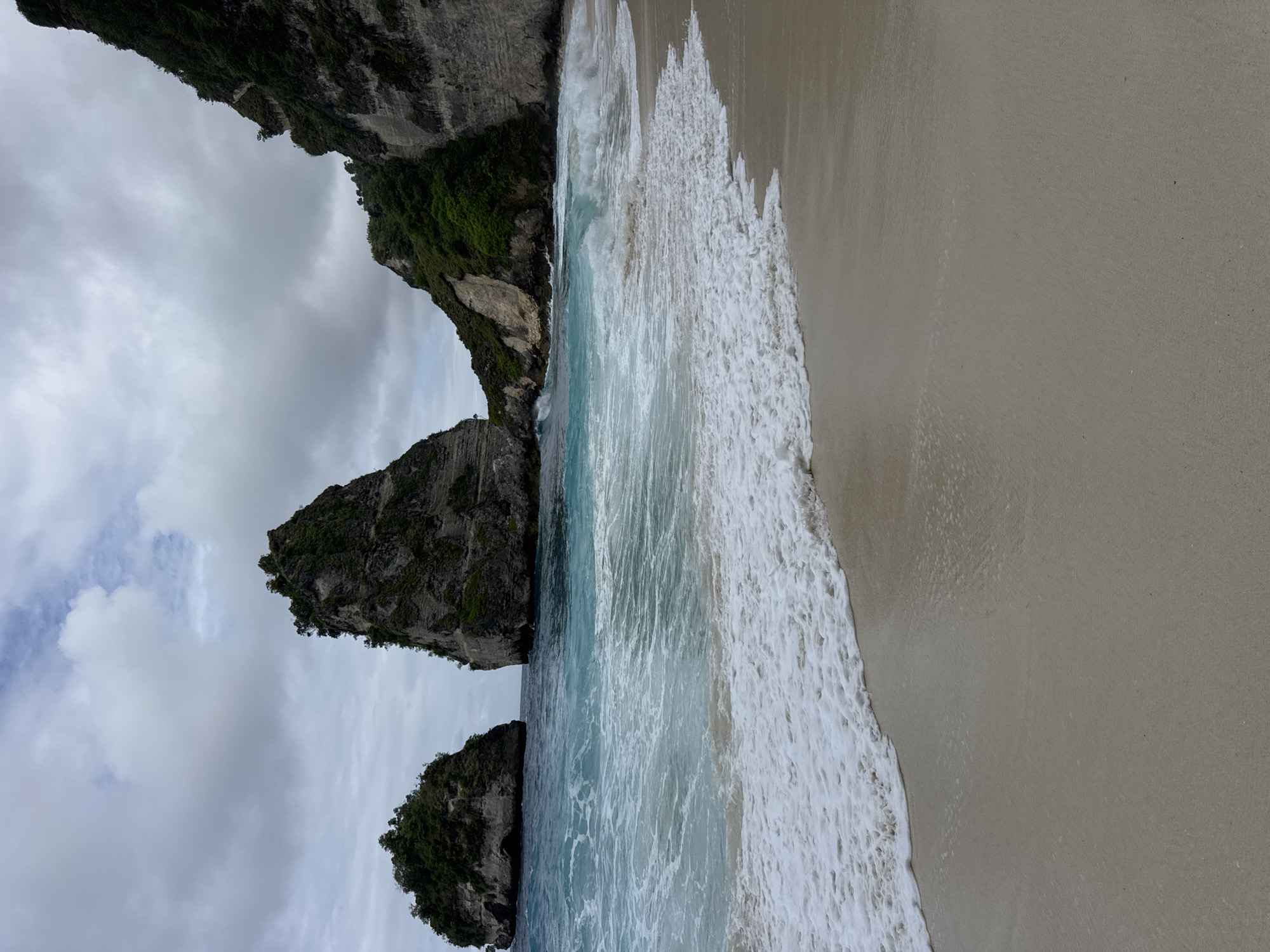 This media file depicts a scenic view of Kelingking Beach on Nusa Penida, Indonesia. The scene is dominated by the iconic cliff formations that resemble a dinosaur's head, covered in lush green vegetation. These dramatic geological features rise steeply from the turquoise ocean.
The activity captured is the gentle lapping of waves onto a sandy beach. The water is a clear, vibrant blue, with white foam from the waves creating a dynamic line along the shoreline. The sand appears light brown and smooth, with some subtle patterns left by the receding water.
The weather appears overcast, with a cloudy sky that diffuses the sunlight. This creates a soft, natural light that highlights the textures of the cliffs and the water. The time of day is not explicitly discernible but the light suggests it could be morning or late afternoon.
Notable details include the sheer scale of the cliffs, the rich greenery clinging to their sides, and the contrast between the rugged rocks and the smooth, inviting beach. The overall impression is one of natural beauty and tranquility. There is no visible text in the image.