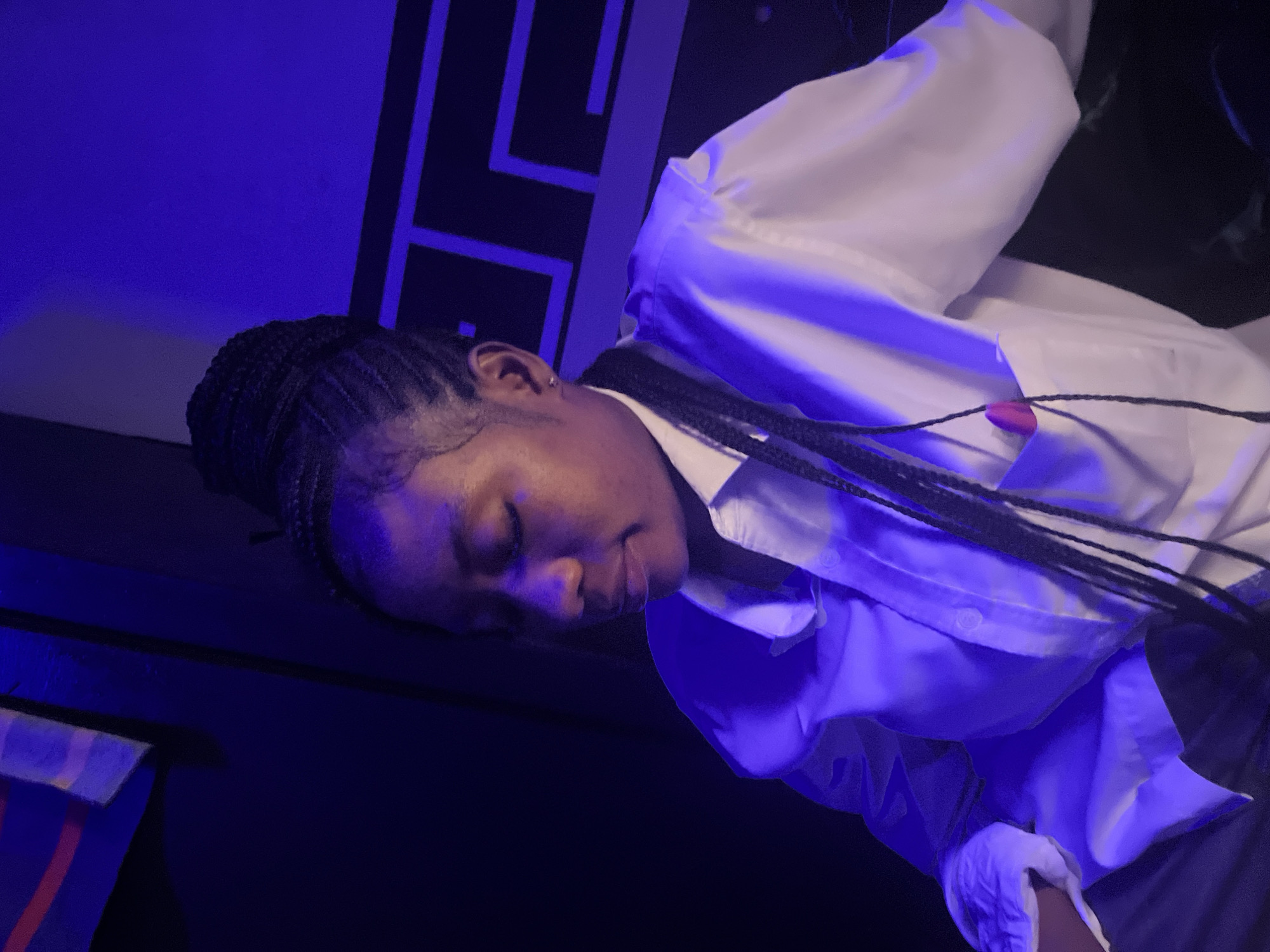 A close-up, rotated shot focuses on the head and upper torso of a young Black woman against a dark background with vibrant blue lighting. She has braided hair, and her eyes are closed with a gentle, contemplative expression. She is wearing a white collared shirt, which is illuminated by the blue light. A black string is visible across the shirt, with a small, pink circular object attached to it. The background features abstract geometric shapes in black and blue. The overall mood is serene and slightly mysterious due to the lighting and the woman's closed eyes. The image appears to be taken indoors, possibly in a studio or performance space, given the controlled lighting and backdrop. There is no visible text.