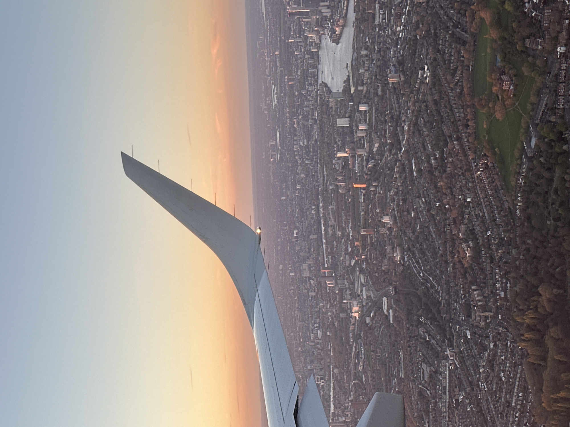 The image shows a view from an airplane window, with the wing of the plane prominently in the foreground against a sky with colors shifting from blue to orange, indicating sunrise or sunset. Below, a densely populated urban area stretches into the distance. Based on the architectural style, arrangement of buildings, and presence of distinct parks, the city is likely London, United Kingdom. The snaking water body is likely the River Thames. Green spaces such as Hampstead Heath are visible in the upper right. The overall impression is that the airplane is either approaching or departing from an airport near London.