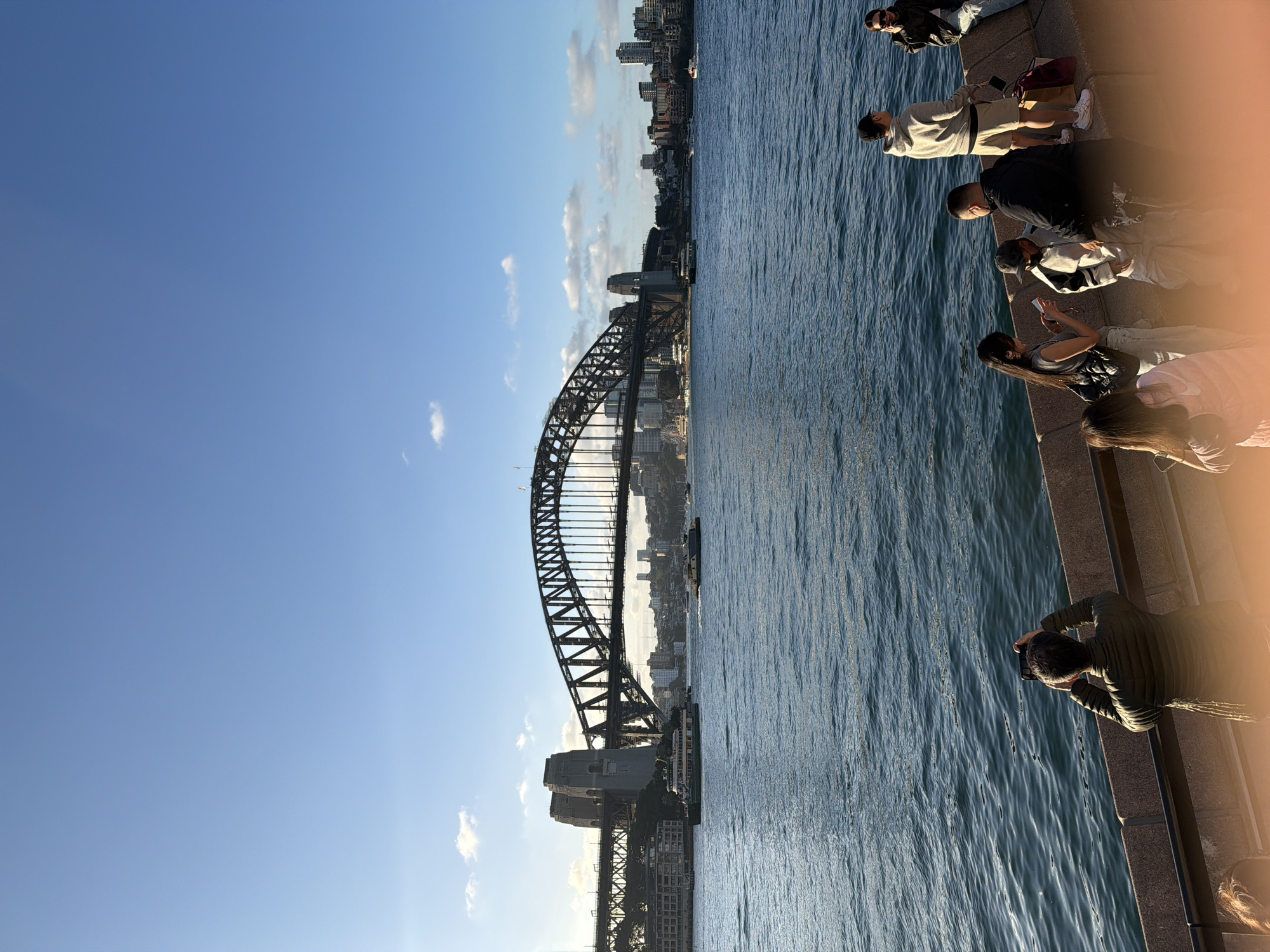 This is a wide-angle, eye-level, outdoor photograph of the Sydney Harbour Bridge in Sydney, Australia. The bridge is the dominant feature, spanning across the frame from left to right, with its iconic arch structure clearly visible. A bright blue sky with scattered white clouds fills the upper portion of the image. The Sydney Harbour, a body of deep blue water, occupies the lower half of the frame, with gentle ripples on its surface.
In the foreground, a concrete embankment runs horizontally, separating the water from a group of people. Several individuals are seated or standing on this embankment, observing the harbour and the bridge. On the left side of the embankment, a person wearing a green puffer jacket is standing and holding up a smartphone, appearing to take a photograph. To their right, a group of people are interacting. One woman is looking at her phone, and a child in a baseball cap is standing next to a man. Another man in a black jacket is seated. Further to the right, a young man in a grey hoodie and shorts is standing, and a woman with sunglasses is seated.
In the middle ground, several ferries are navigating the harbour. The largest ferry, with multiple decks and windows, is moving from left to right. Other smaller boats and vessels are also visible on the water. The cityscape of Sydney is visible in the background, with buildings and skyscrapers dotting the horizon behind the bridge. The sun appears to be setting or rising, casting a warm light on the scene.