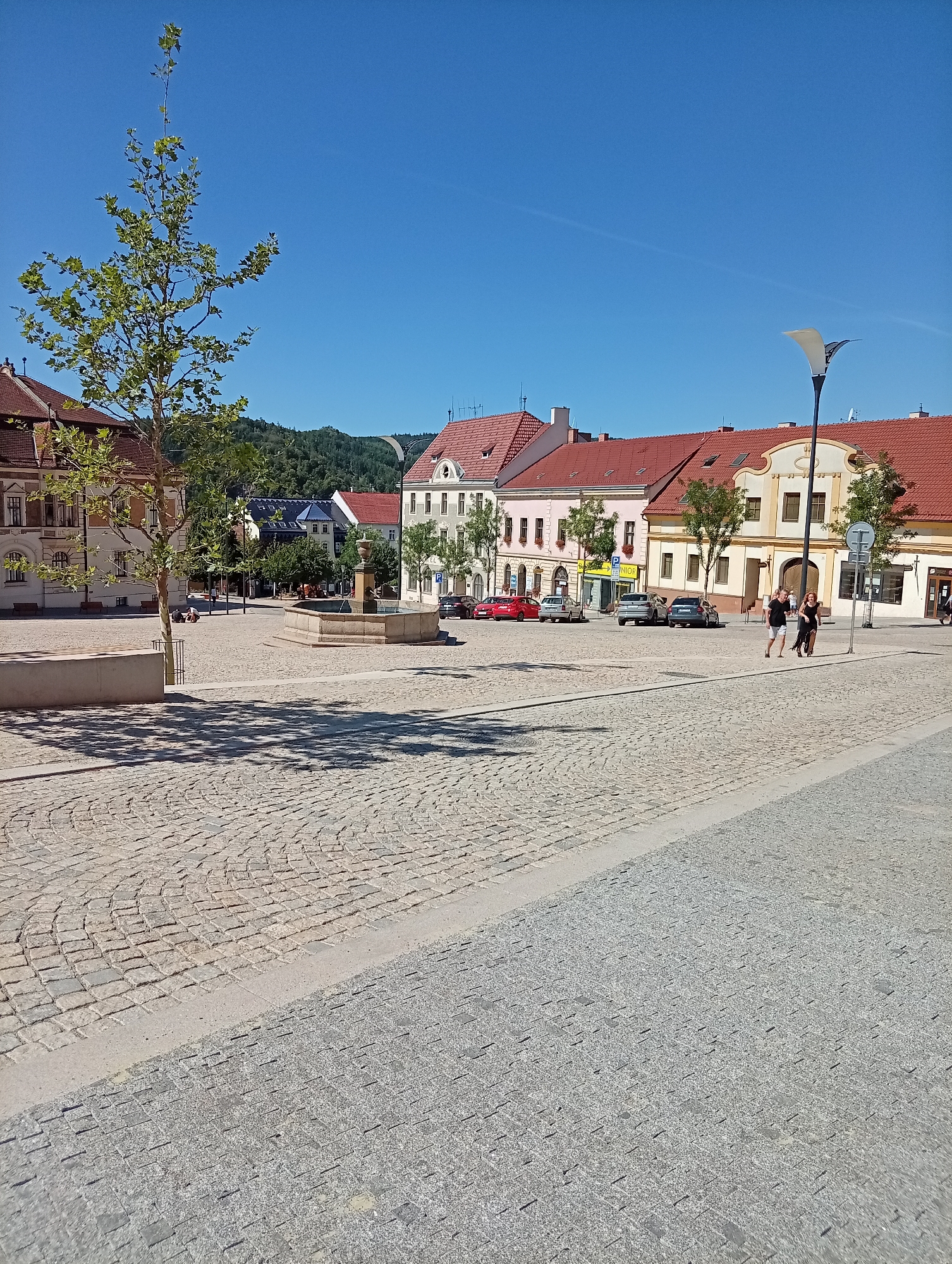 The image depicts a sunlit public square in Tišnov, Czechia. The square is paved with cobblestones, featuring a low-lying stone bench on the left and a stone fountain in the mid-ground. Several trees are scattered throughout the square, including a young tree in the left foreground casting a dappled shadow.
In the background, a row of buildings with red tiled roofs line the square. These buildings exhibit traditional European architecture with multiple windows and decorative facades. Some of the buildings appear to house shops, indicated by signage. Several cars are parked along the street in front of the buildings.
Two individuals are walking across the square in the mid-right of the frame. One is wearing a black dress, and the other is in dark shorts and a t-shirt. A modern lamppost stands to the right of the individuals. The sky is a clear, vibrant blue, suggesting fair weather. In the distance, a forested hill is visible.