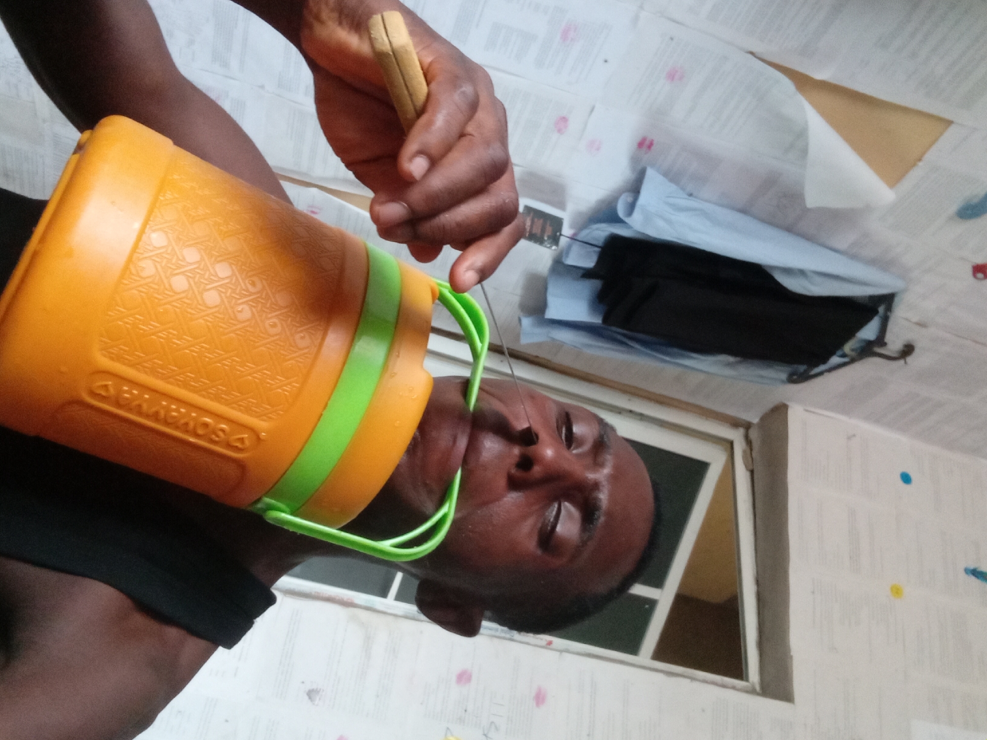 A man is shown holding an orange and green container to his face. The container appears to be a water carrier or cooler of some kind. He is holding what appears to be a small, thin object, possibly a needle or pin, near his nose. The location appears to be indoors, possibly a bedroom, with a window visible in the background and a pile of clothes on the floor. The location is Unknown, Unknown. The man's expression and the action of holding the object near his nose suggest a possible extraction or procedure.