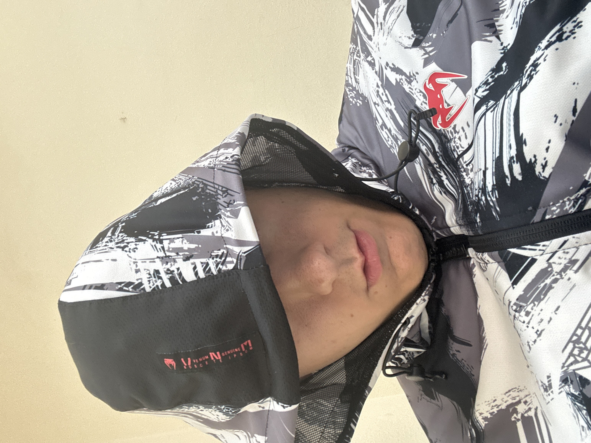 The image is a close-up, angled shot of a person wearing a hooded jacket, presumably indoors in Plovdiv, Bulgaria. The person's face is partially visible, showing their nose, mouth with pink lips, and chin, framed by the hood of the jacket.
The jacket has a distinctive black, white, and grey abstract pattern, resembling brushstrokes or grunge art. A prominent red logo, appearing as a stylized letter or a horned figure, is visible on the right chest area of the jacket. The hood features a black mesh lining and a black brim. Along the brim of the hood, there is visible text in white and red, which reads "VENUM N GENUINE M HORCCE T", identifying a brand. The jacket also includes black drawstrings with toggles.
The background is a plain, light beige or cream-colored wall. The unusual angle of the shot causes the person's head to appear tilted sharply to the left. No specific activity is discernible beyond the person wearing the jacket. The indoor setting does not provide clues about the time of day or weather.