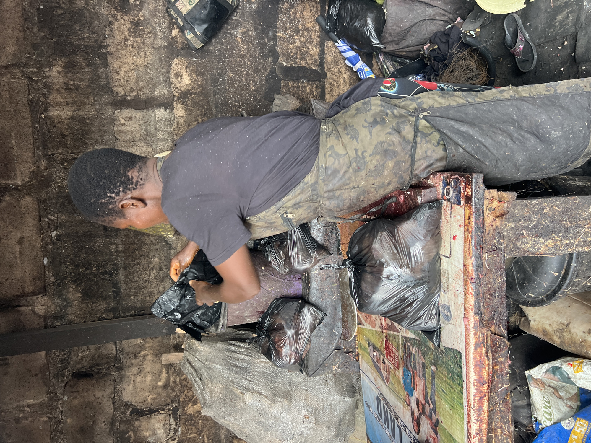 The image depicts a woman wearing a black t-shirt and a camouflage apron, standing behind a wooden counter in what appears to be an outdoor market stall in Nkpor, Nigeria. The stall is cluttered with various items, including several black plastic bags filled with goods, a large grey sack, and a purple bucket. The woman is in the process of handling a black plastic bag, suggesting she is either packing or unpacking items. The background consists of a rough-textured wall and a metal beam, indicating a rustic and possibly unfinished structure. The overall environment appears to be a working space, likely involved in the sale of goods.