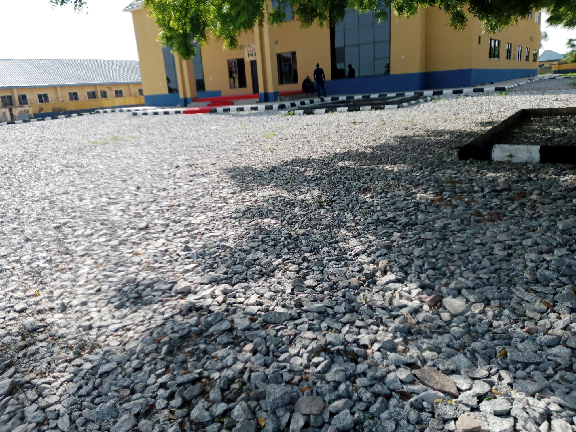The image captures an outdoor scene featuring a two-story building with a yellow facade and a blue base, situated in Jalingo, Nigeria. In front of the building, there is a gravel-covered area. Several individuals are present on the steps leading up to the building. One person is standing, dressed in a dark blue shirt and trousers, while another individual is seated nearby, also in dark clothing. Two other figures are visible further back, appearing to be inside the building or near its entrance. The building's signage indicates it is the "INSTITUTE OF CONST. NI". To the left of the main building, a longer, single-story structure with a grey corrugated roof is partially visible. The foreground is dominated by the textured surface of the gravel. The scene appears to be an educational or administrative complex.