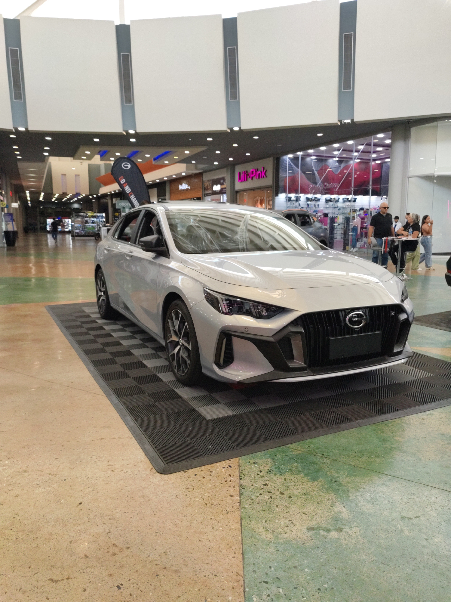 A silver sedan is displayed indoors, likely at a mall or exhibition center, on a checkered black mat. The car is positioned at an angle, showcasing its front grille, headlights, and side profile. In the background, several shops are visible, including one with a neon pink sign that reads "Lili-Pink." A few people are standing and interacting near a counter in the background. The flooring appears to be a patterned material with green and tan sections. The overall environment suggests a commercial or public space, potentially in the city of Palo Negro, Venezuela, given the context.