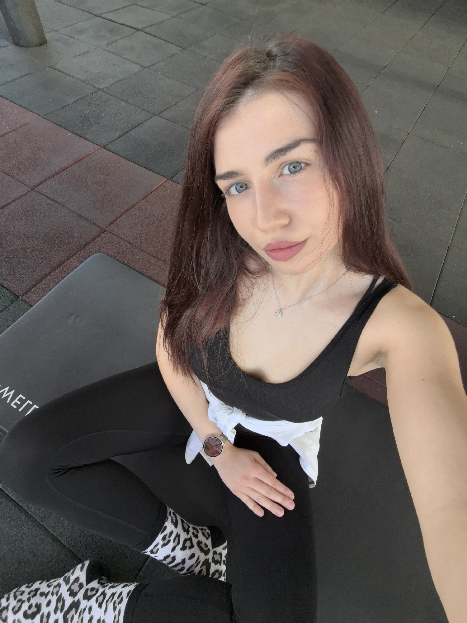 A young woman is seated on a black exercise mat. She wears black leggings, a black tank top, and leopard-print socks. A white shirt is tied around her waist. The woman has long, dark brown hair and blue eyes. She wears a necklace with a heart pendant and a watch on her left wrist. The flooring consists of interlocking black and red rubber tiles. The photo seems to have been taken indoors. This information does not provide enough context to determine whether it was taken in Stara Zagora.