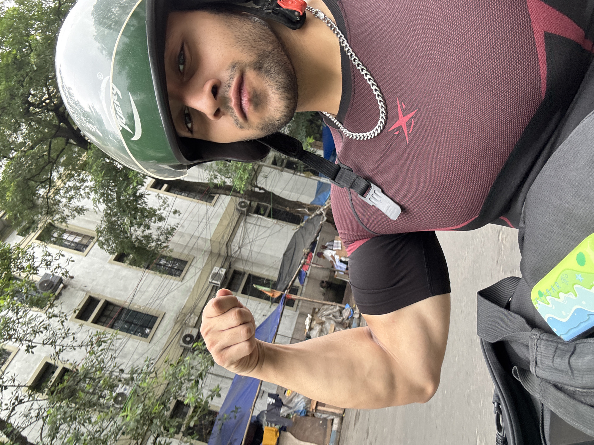 The image shows a man in a helmet flexing his bicep. He is wearing a maroon and black compression shirt with a red emblem on it and a silver chain necklace. He appears to be on a street, possibly near a building with multiple windows and air conditioning units. In the background, there are some makeshift structures covered with blue and black tarpaulins, suggesting a temporary settlement or market. There are also trees and electrical wires visible, consistent with an urban environment. The man's pose and focus suggest he is taking a selfie. The scene is likely in Kolkata, India, given the architectural style of the building and the presence of the makeshift shelters, which are common in parts of the city.