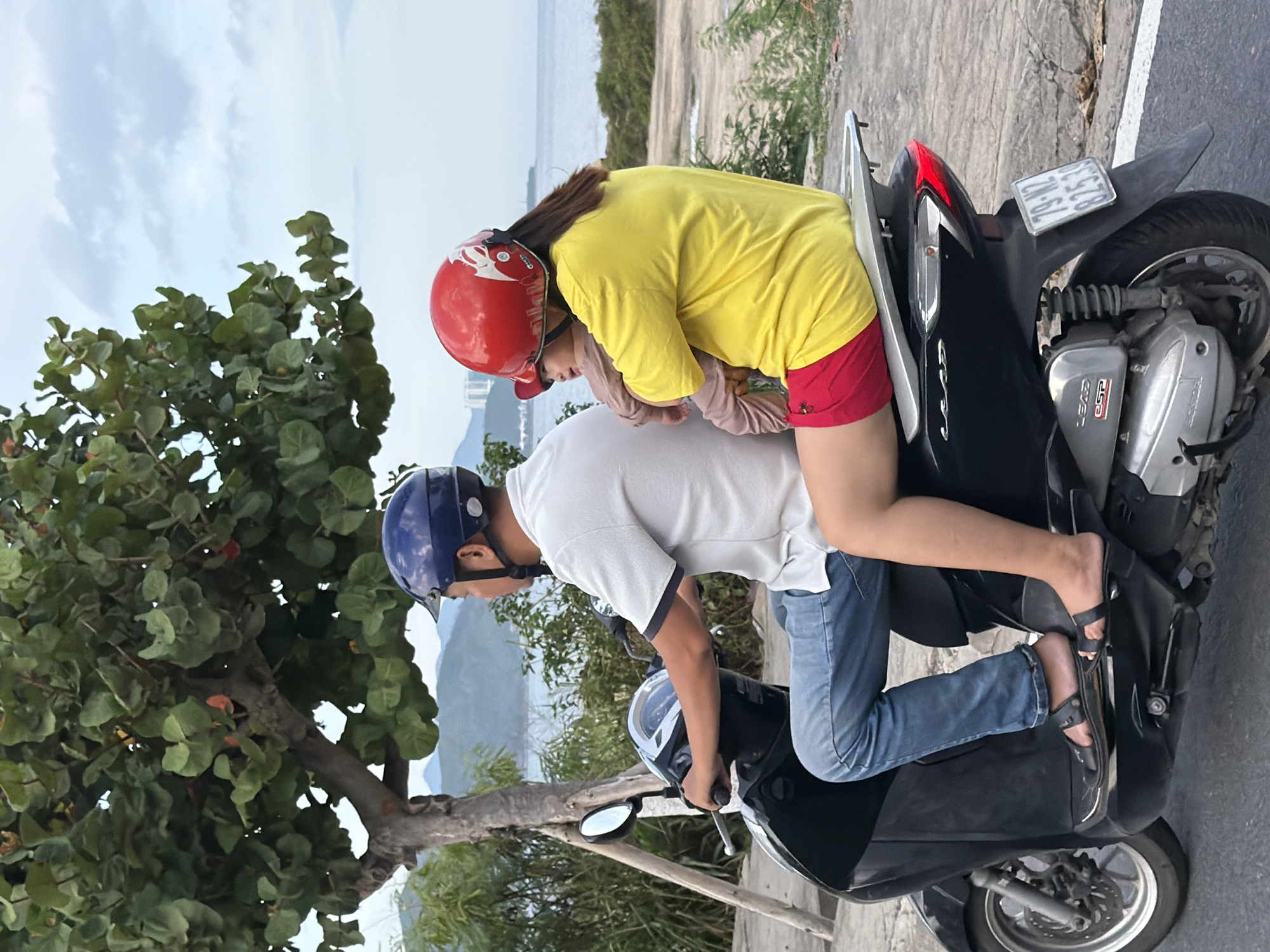 The image captures a family of three riding a scooter on a paved road in Nha Trang, Vietnam. The father, wearing a blue helmet, is driving the scooter and has a white polo shirt and blue jeans on. The mother, with a red helmet, is seated behind him, holding a baby. She is wearing a yellow t-shirt and red shorts. The baby appears to be wearing light brown clothing. The background shows lush green trees, a body of water, and distant hills under a cloudy sky. The scooter is black and has a license plate that reads "79-N2 825.39". The overall scene depicts a typical mode of transportation in Vietnam, with a focus on the everyday life of a family.
